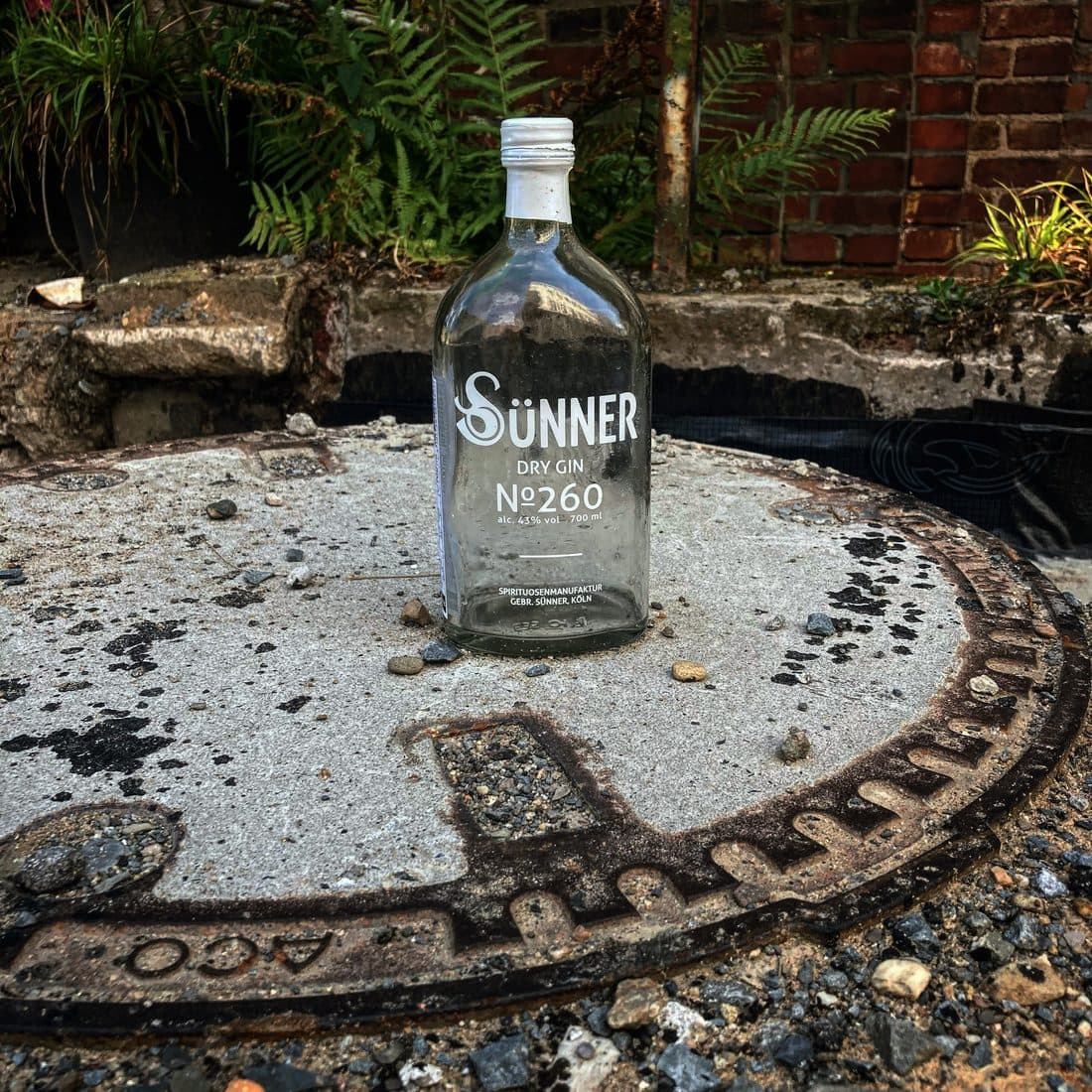 Eine leere Flasche Sünner Dry Gin steht auf einem runden Schachtdeckel aus Beton mit rostigem Eisenrand, dahinter bröckelnder Beton, Farne und eine rote Backsteinmauer