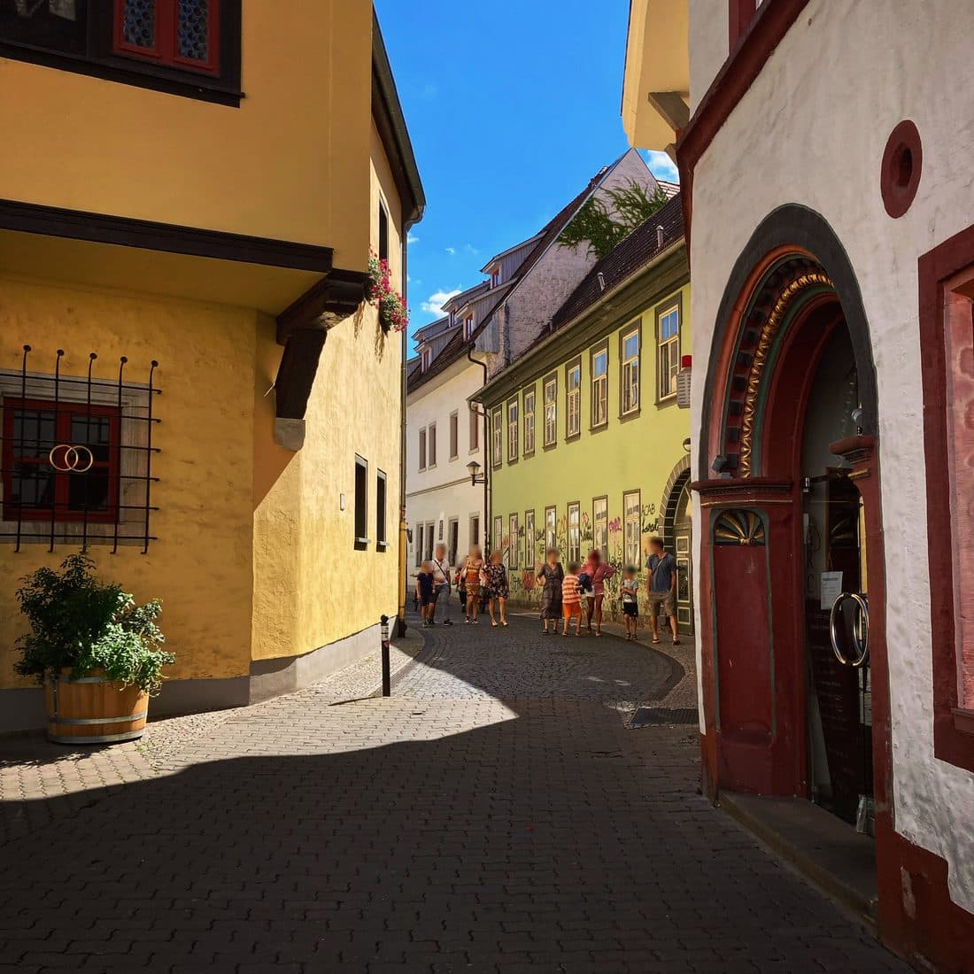Eine Altstadtgasse mit Pflaster im Sonnenlicht, links ein gelbes Haus, rechts ein weißes mit rotem Rundbogenportal, weiter hinten laufen mehrere Menschen vor einer hellgrünen Fassade