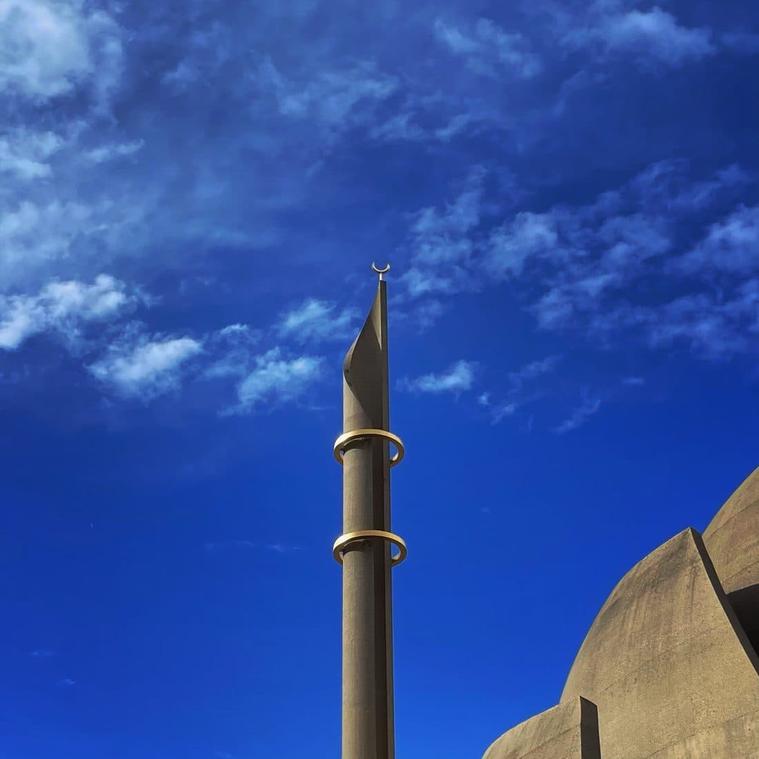 Das schlanke Betonminarett einer Moschee mit zwei goldenen Ringen und einem Halbmond an der Spitze vor tiefblauem Himmel mit lockeren Wolken, rechts unten die Rundung einer Betonkuppel