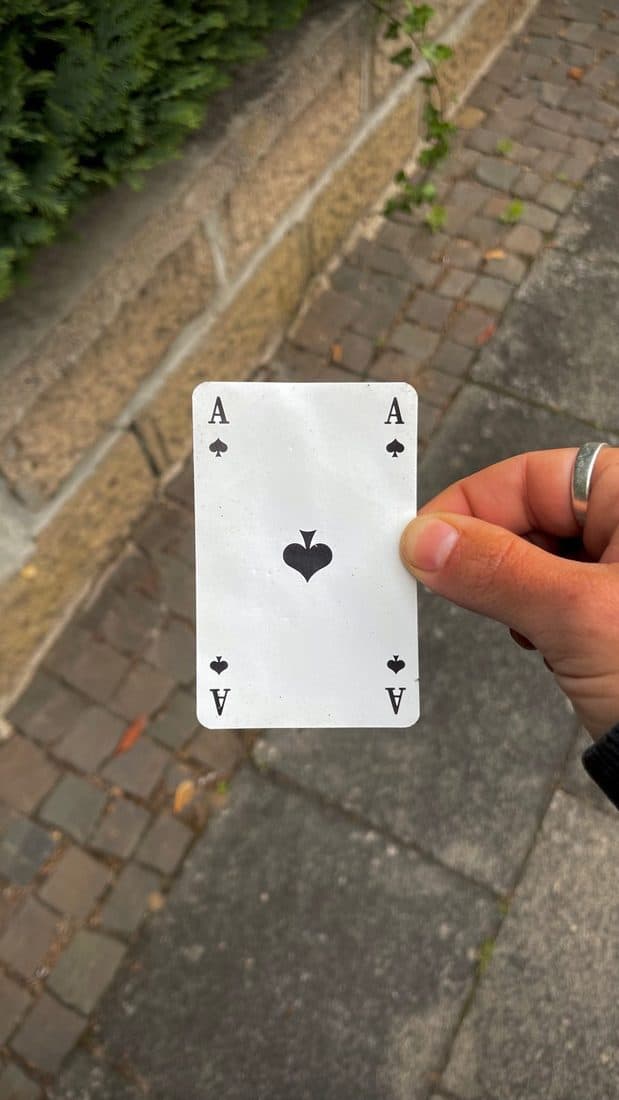 Eine Hand hält ein Pik-Ass aus einem Kartenspiel über einen Weg aus Kopfsteinpflaster