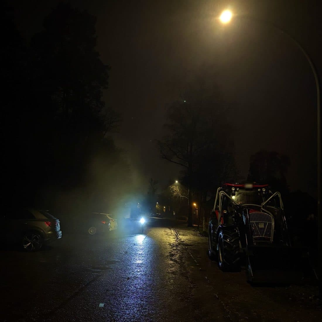 Nächtliche Straße im Nebel: eine Laterne leuchtet warm in den Dunst, links parkende Autos, rechts eine rote Forstmaschine, der nasse Asphalt spiegelt das Licht