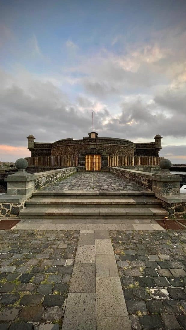 Ein rundes Steinkastell mit hölzernem Tor am Meer, davor ein gepflasterter Weg mit Stufen, am Himmel Abendwolken