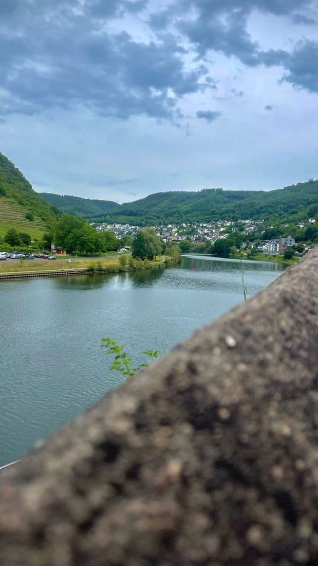 Blick über einen ruhigen Fluss auf ein Dorf zwischen bewaldeten Hügeln und Weinbergen, im Vordergrund unscharf eine Mauerbrüstung