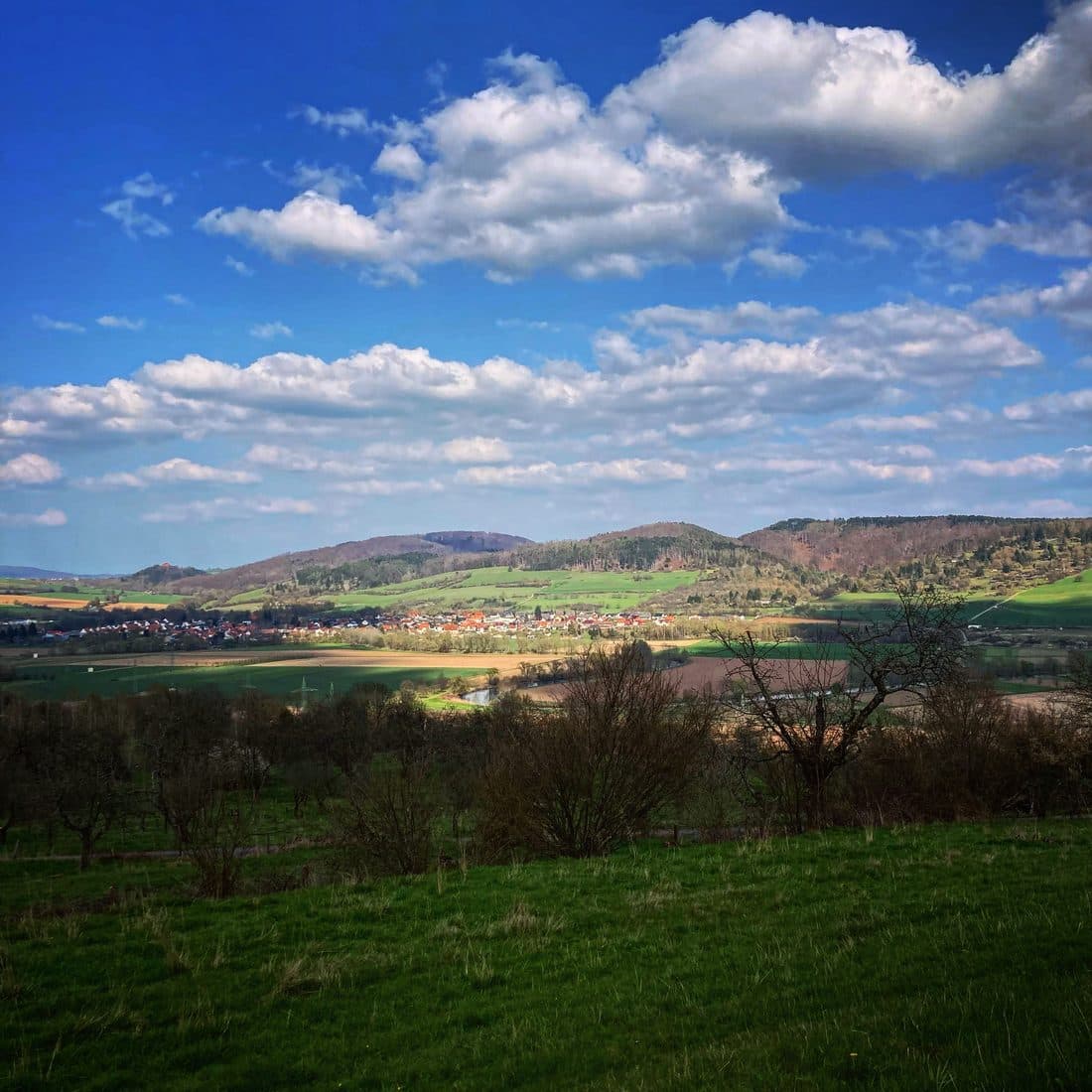 Ein Dorf mit roten Dächern in einem Tal vor bewaldeten Hügeln, im Vordergrund eine grüne Wiese und kahle Bäume, darüber tiefblauer Himmel mit weißen Haufenwolken
