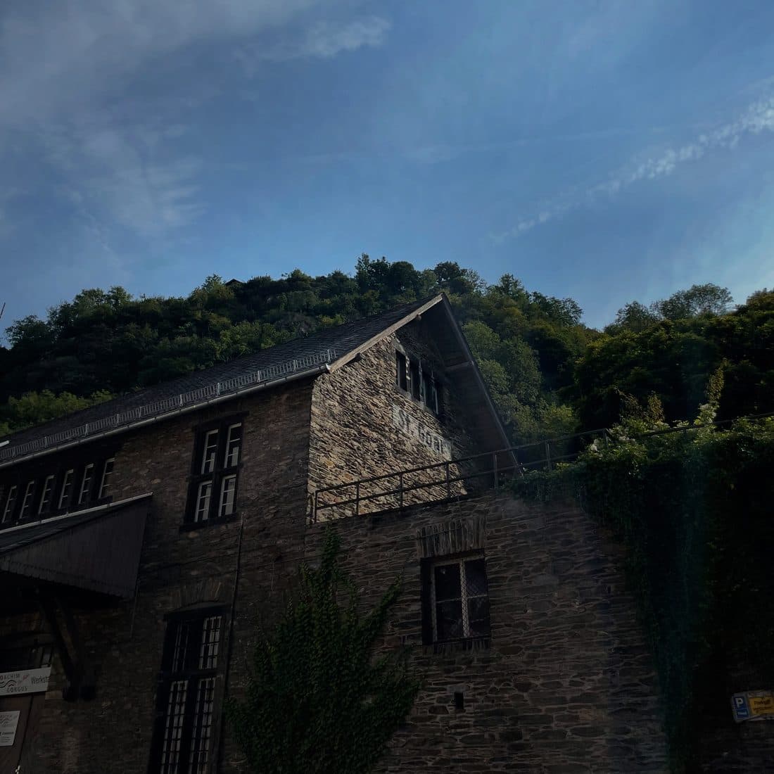 Ein altes Bruchsteinhaus mit verblasstem Schriftzug St. Goar am Giebel liegt im Schatten vor einem bewaldeten Hang, darüber blauer Himmel mit zarten Wolken