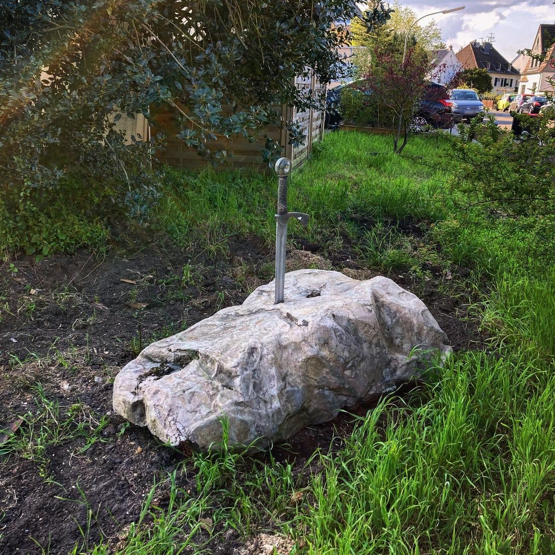 Ein großer heller Stein mit einem darin steckenden Schwert liegt auf dunkler Erde zwischen hohem Gras, dahinter Sträucher, ein Holzzaun, parkende Autos und Wohnhäuser