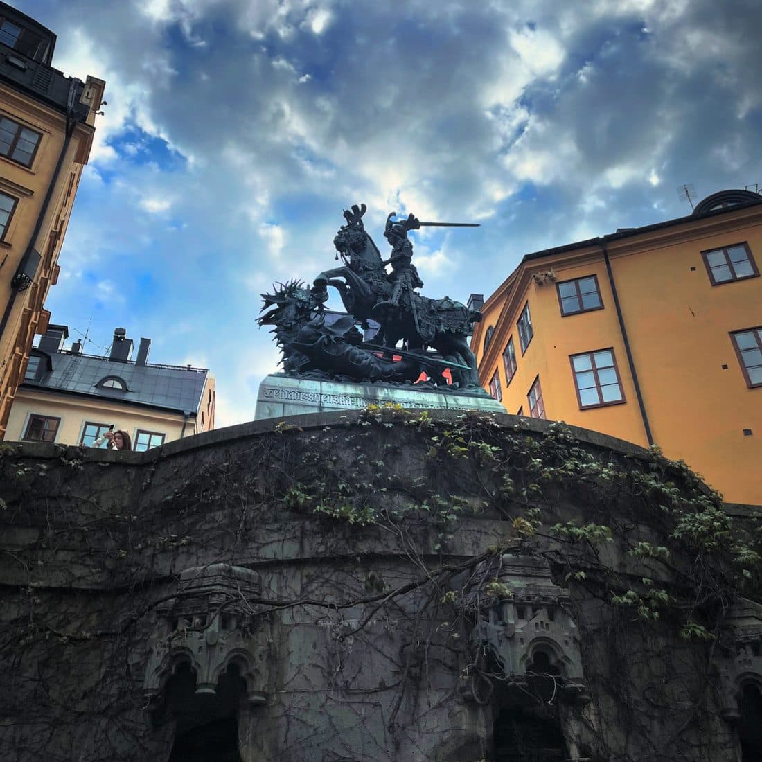 Das bronzene Reiterstandbild des heiligen Georg mit dem Drachen in Stockholm ragt über einer rankenbewachsenen Steinmauer auf, umgeben von ockergelben Häusern und Wolken