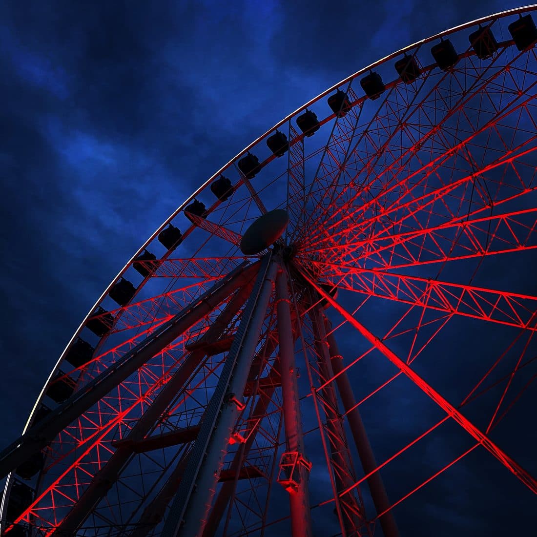 Ein Riesenrad von unten, sein Stahlgerüst rot angestrahlt, die dunklen Gondeln am äußeren Ring vor einem tiefblauen, bewölkten Abendhimmel