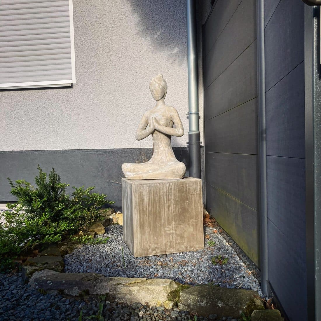 Eine helle Steinfigur sitzt im Schneidersitz mit vor der Brust aneinandergelegten Händen auf einem Sockel im Kiesbeet, neben einer Hauswand mit Rollladen und dunkelgrauen Paneelen