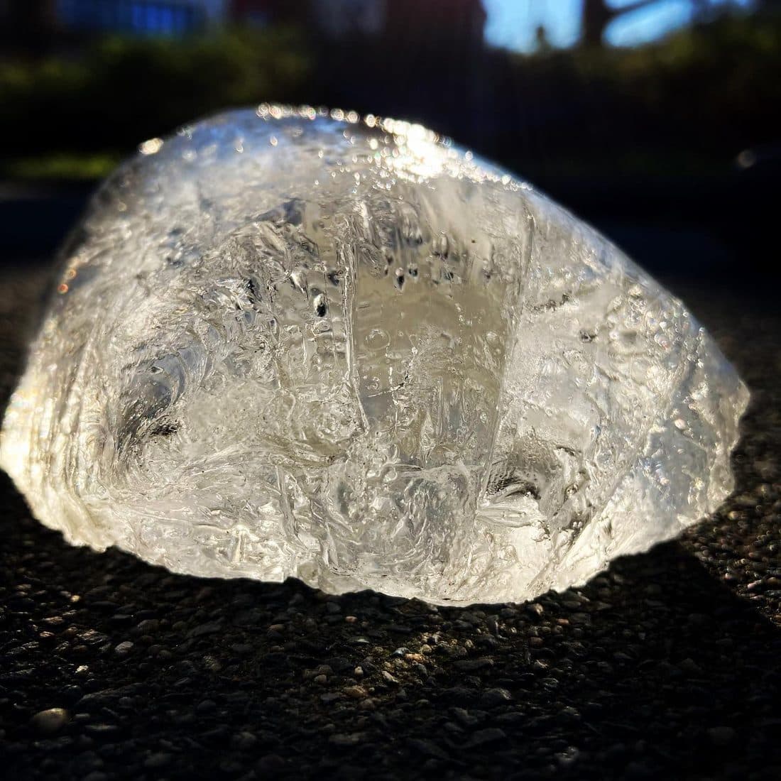 Ein Stück klares Eis mit eingeschlossenen Luftblasen liegt auf dunklem Asphalt und leuchtet im Gegenlicht der tief stehenden Sonne, dahinter dunkle, unscharfe Bäume