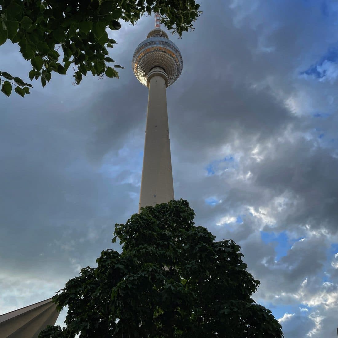 Der Berliner Fernsehturm von unten gesehen, seine Kugel im warmen Licht vor grauen Wolken mit blauen Lücken, unten ragt eine dunkle Baumkrone ins Bild