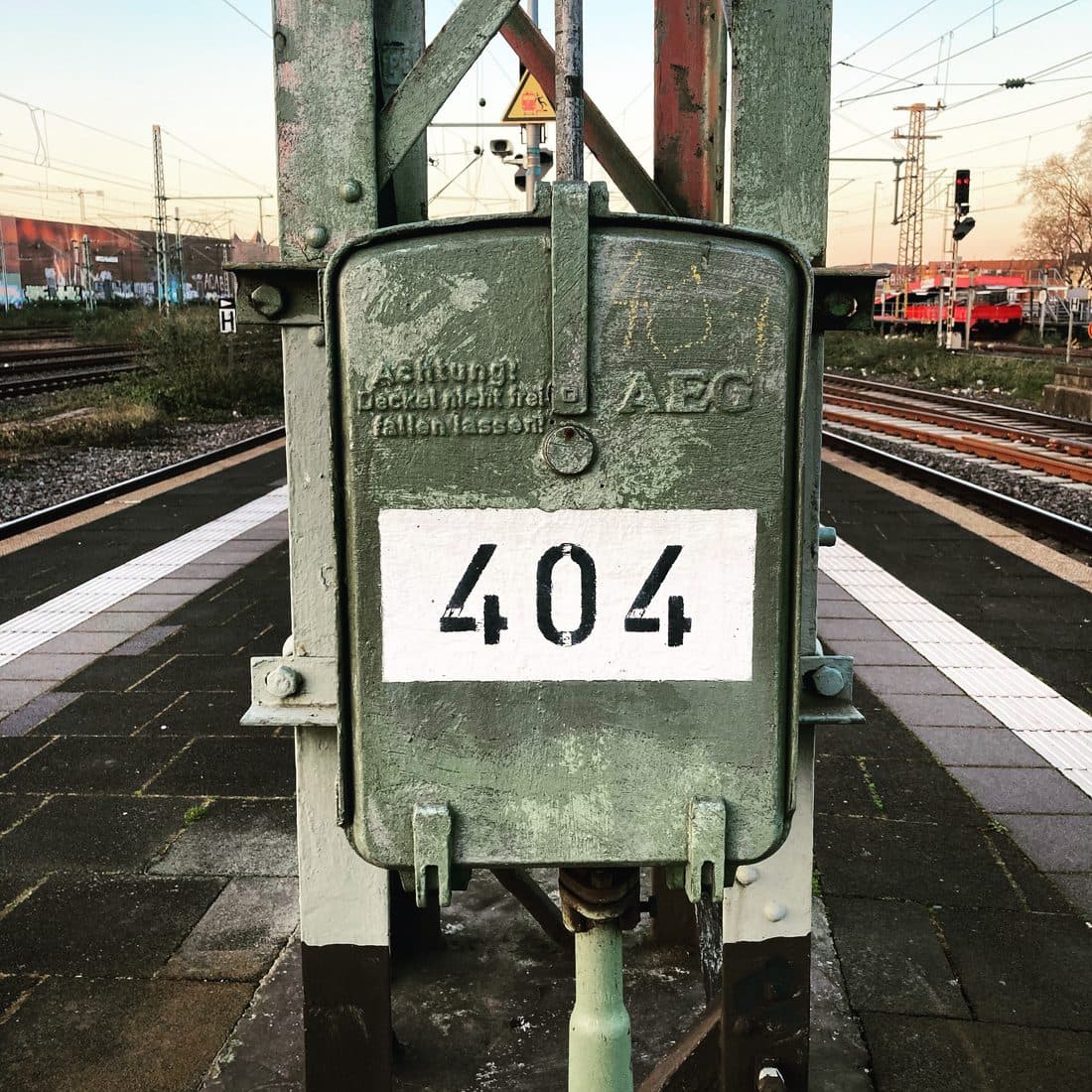 Ein grün gestrichener Metallkasten mit der schwarzen Zahl 404 auf weißem Feld an einem Mast auf einem Bahnsteig, zu beiden Seiten Gleise und Oberleitungen im Abendlicht