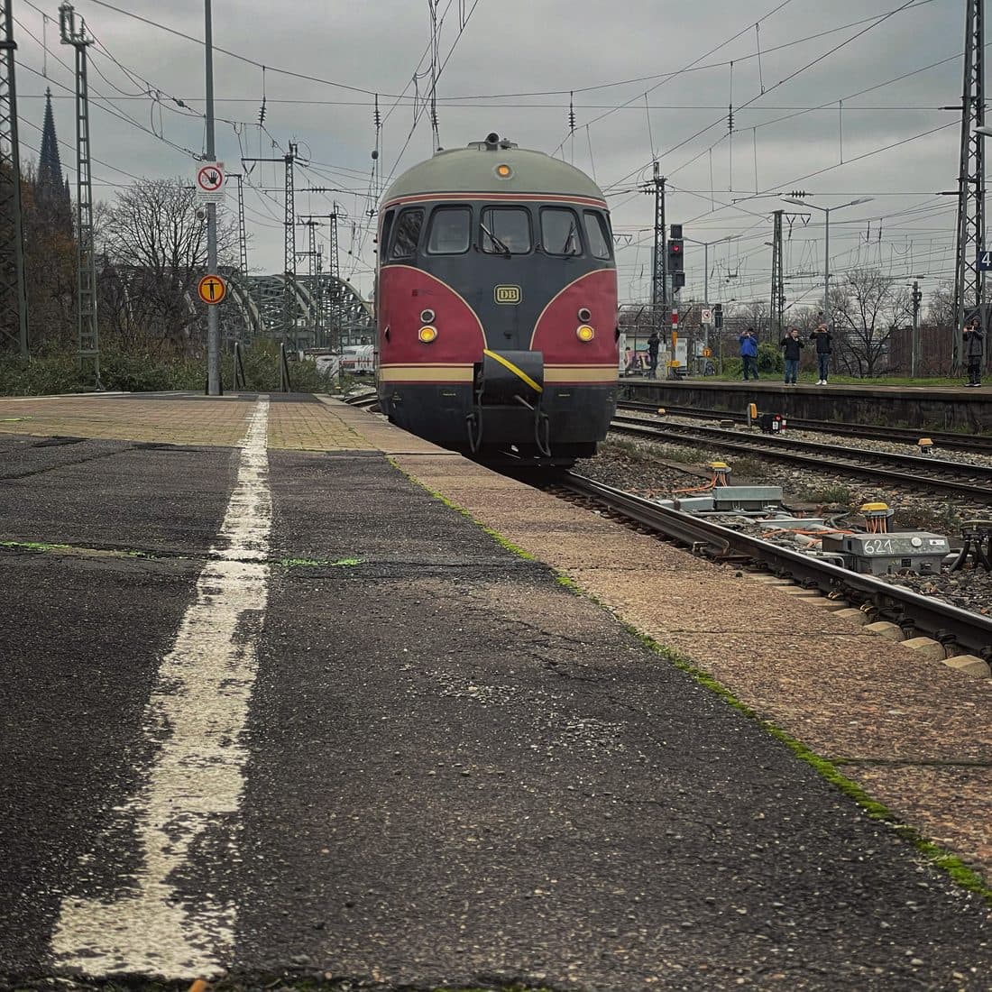 Ein historischer Triebwagen in Weinrot und Dunkelgrau neben einem leeren Bahnsteig, links hinten ein Turm des Kölner Doms und die Hohenzollernbrücke, rechts fotografieren Menschen den Zug