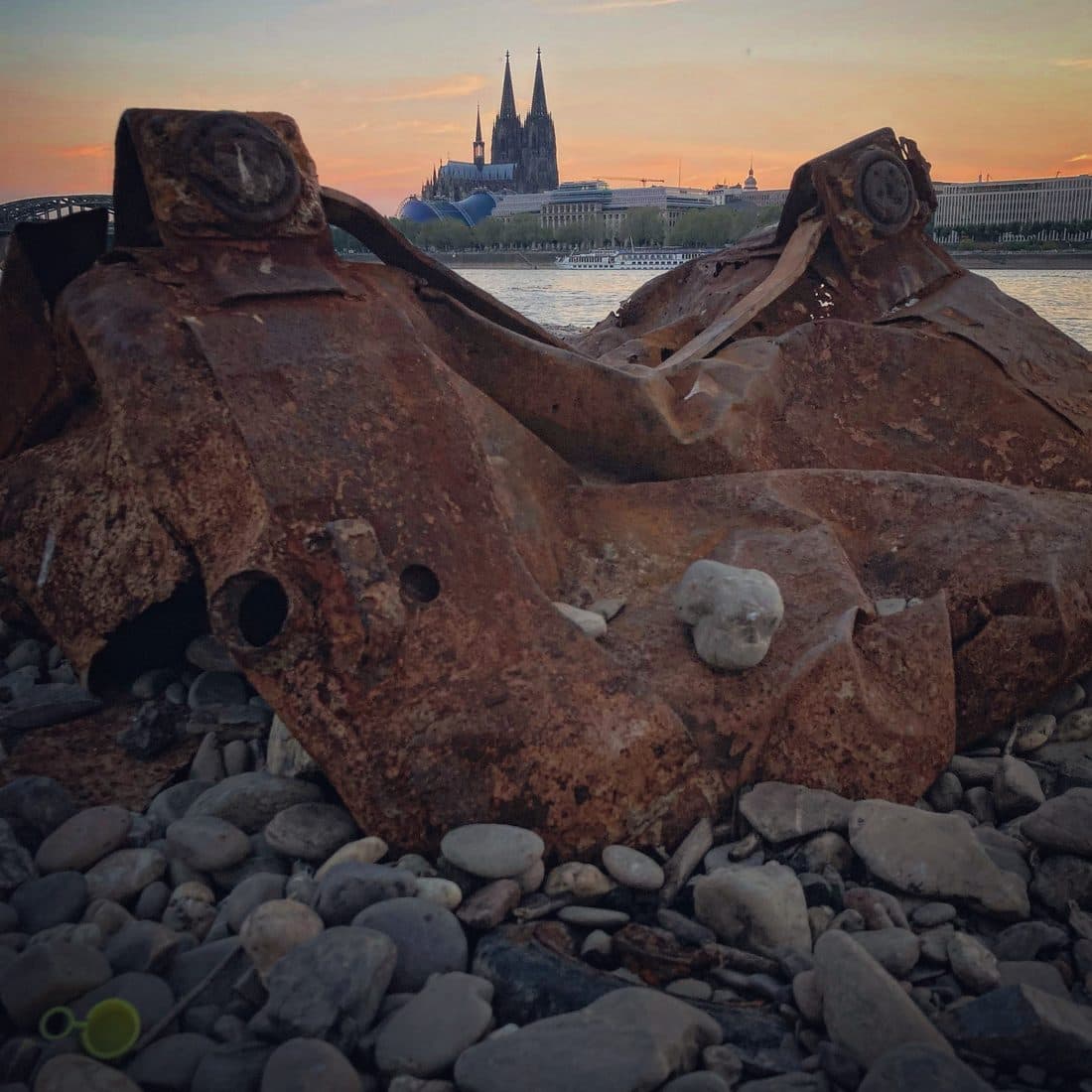 Ein stark verrostetes, verbeultes Metallteil liegt auf Kieselsteinen am Rheinufer, auf der anderen Flussseite ragen die Türme des Kölner Doms in den orangefarbenen Abendhimmel