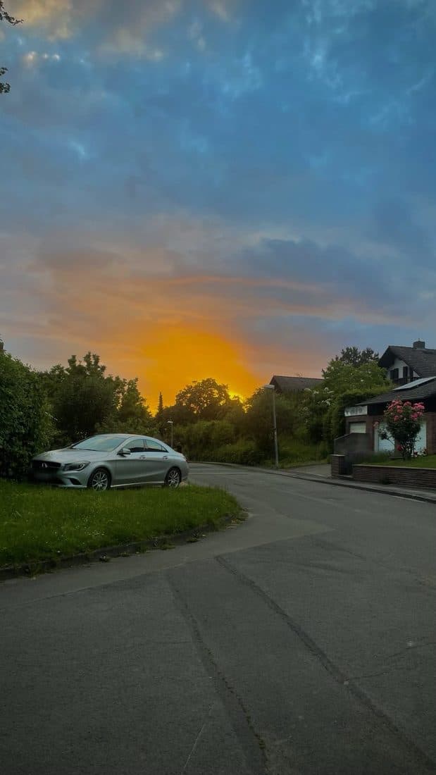 Eine leere Wohnstraße am Abend, über den Bäumen glüht der Himmel orange, links parkt ein silbernes Auto im Gras