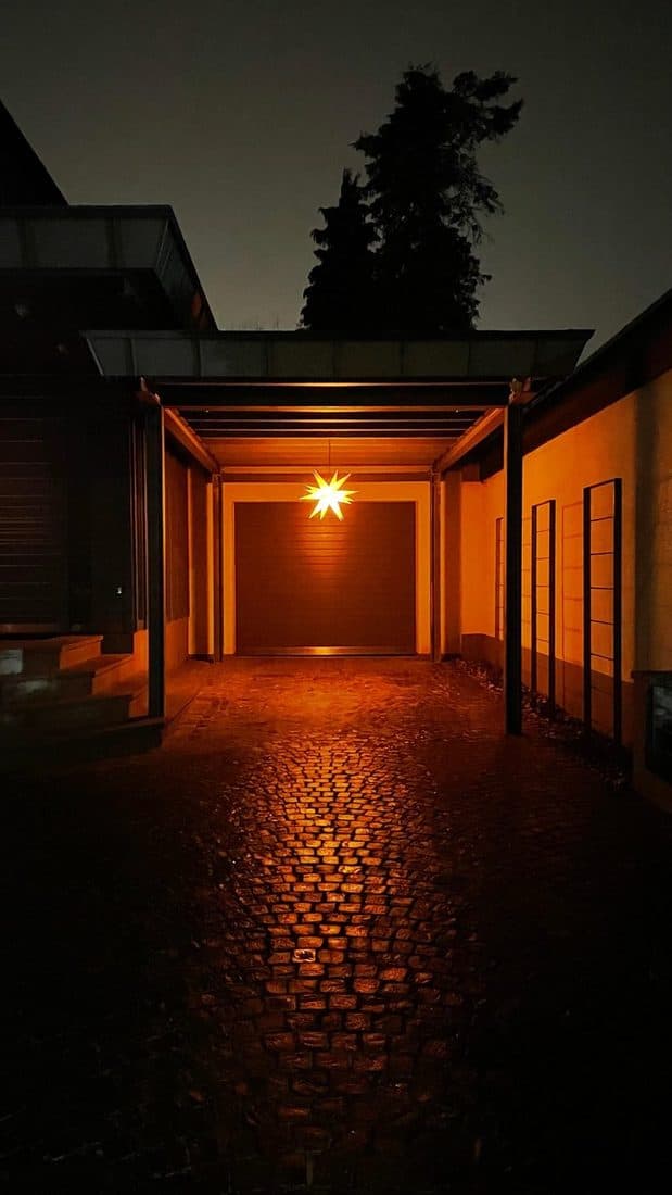 Nachts leuchtet ein gelber Papierstern unter einem Vordach und wirft warmes Licht auf das nasse Kopfsteinpflaster