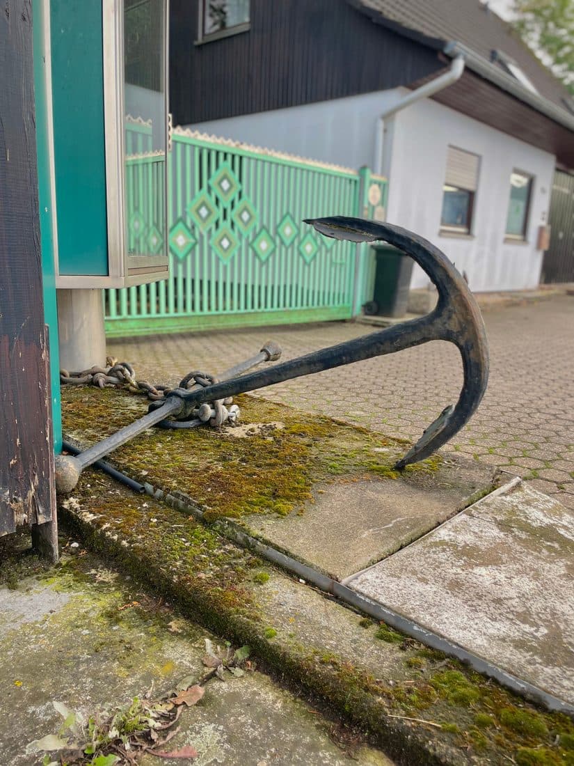Ein schwarzer Schiffsanker mit Kette liegt auf einer bemoosten Betonplatte vor einem Haus, dahinter ein mintgrünes Tor mit Rautenmuster und eine gepflasterte Einfahrt