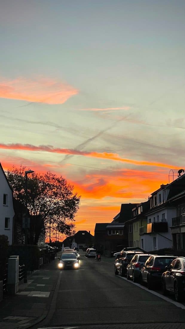 Eine Wohnstraße in der Dämmerung, über den Dächern glühen orange Wolkenbänder, ein Auto kommt mit eingeschaltetem Licht entgegen