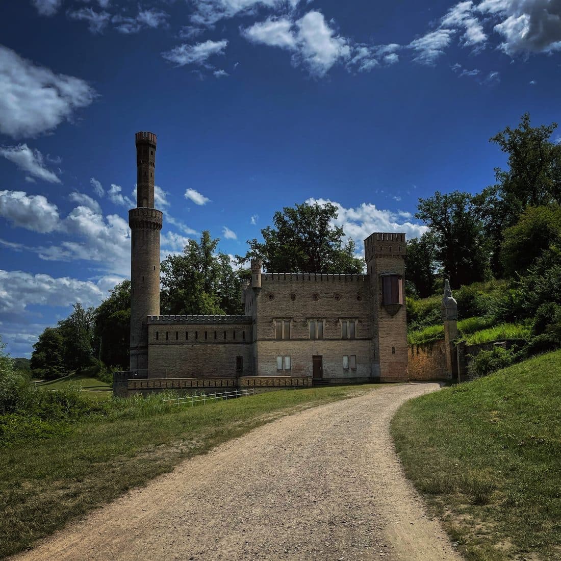 Ein burgartiges Gebäude aus gelblichem Backstein mit Zinnen und einem hohen, schlanken Rundturm, davor ein geschwungener Kiesweg, ringsum Bäume unter tiefblauem Himmel