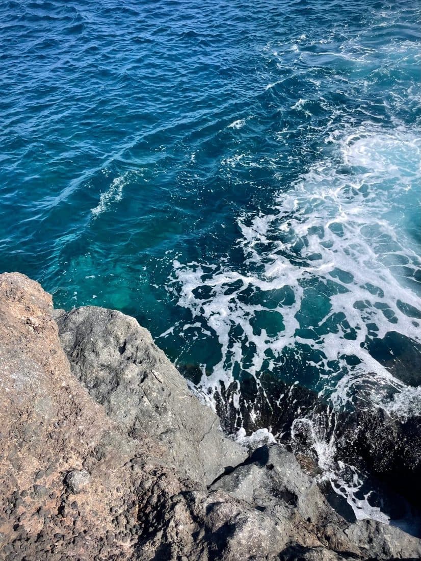 Von oben auf tiefblaues Meer: weiße Schaumfäden ziehen über das Wasser, unten links ragen graue Lavafelsen ins Bild
