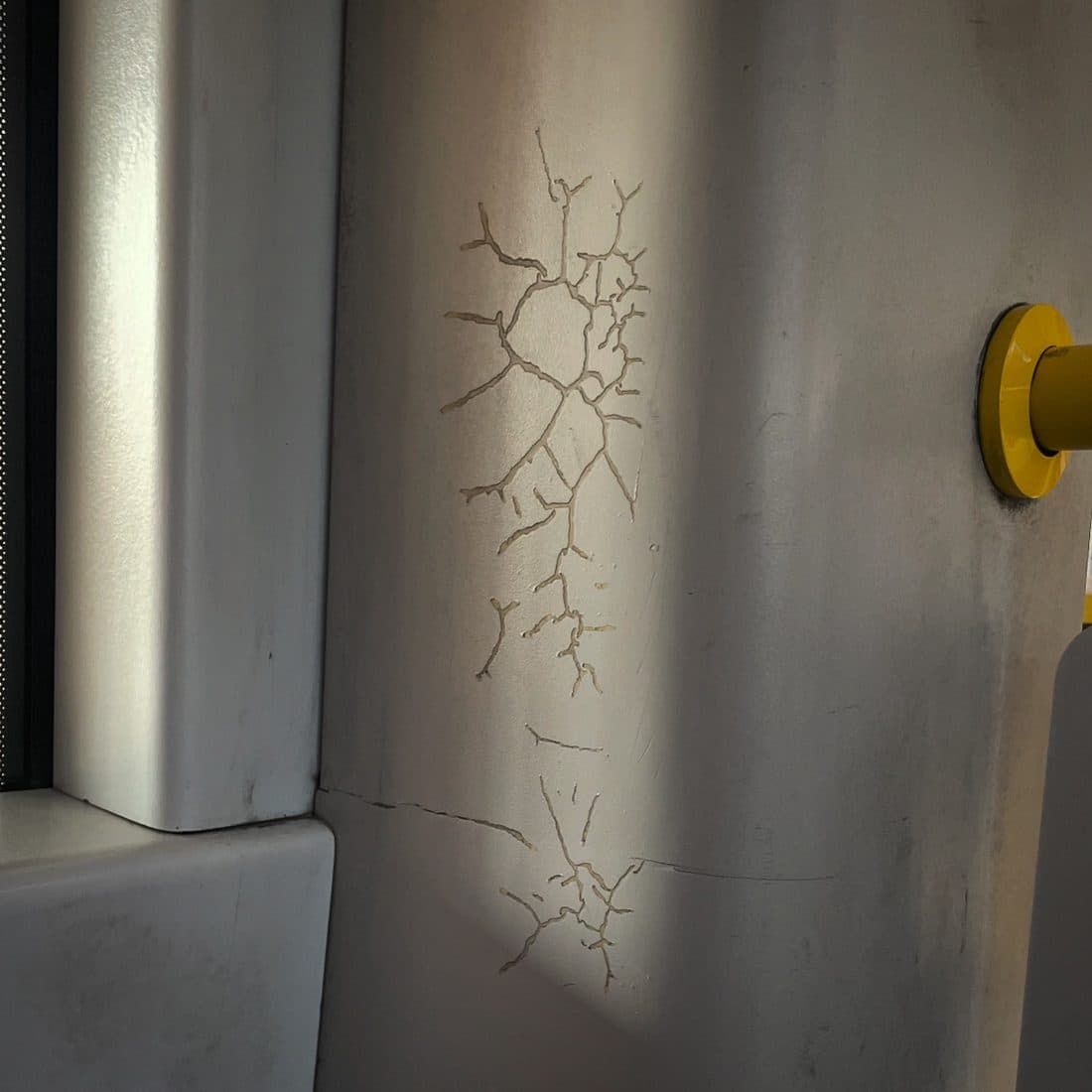 Ein verzweigtes Rissmuster in einer hellgrauen Verkleidung neben einem Fensterrahmen, rechts ein gelber Haltegriff, darüber ein schräger Streifen Sonnenlicht