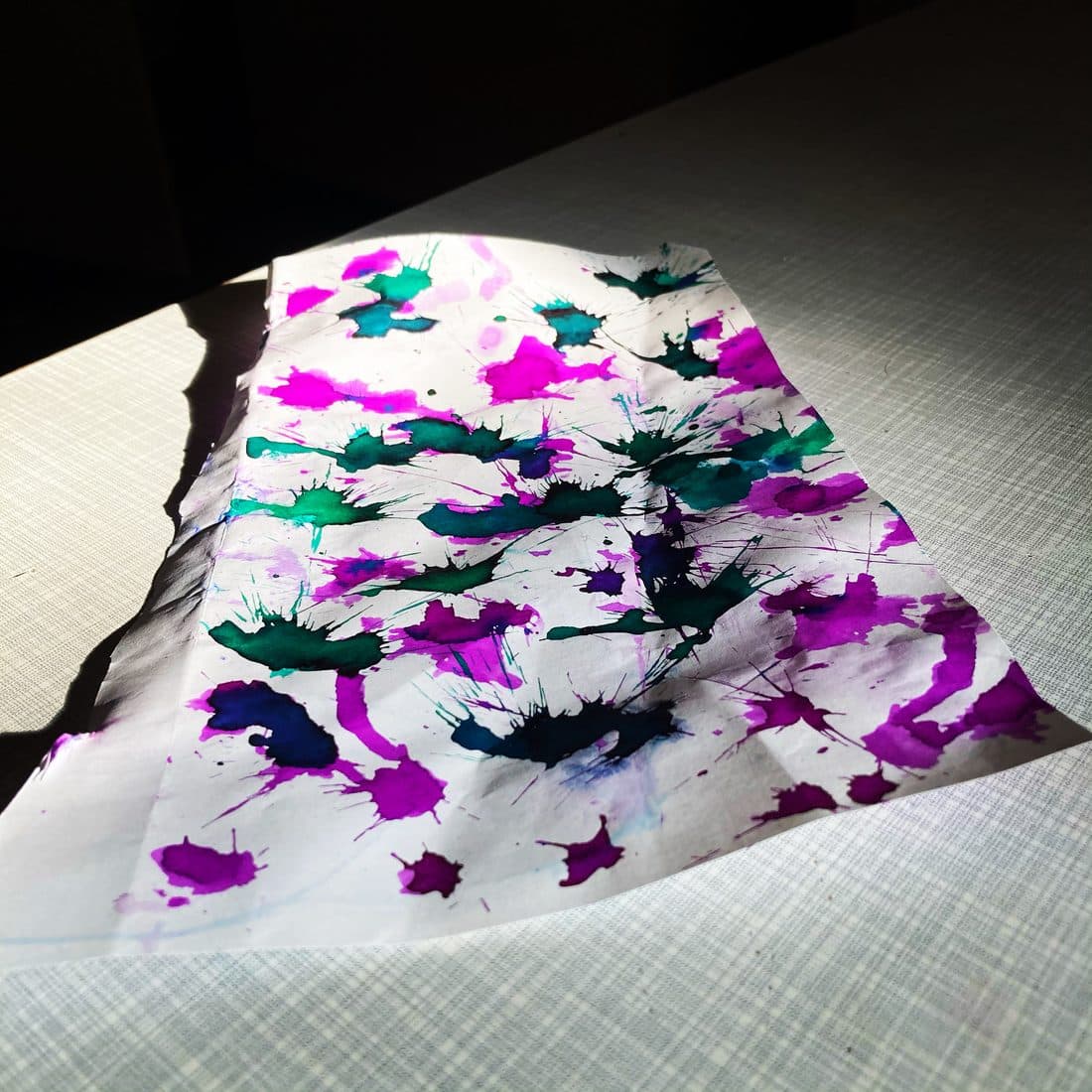 Ein weißes, leicht gewelltes Blatt Papier mit Klecksen und Spritzern in Magenta, Dunkelgrün und Blau liegt im schrägen Sonnenlicht auf einer hellen, fein gemusterten Tischplatte