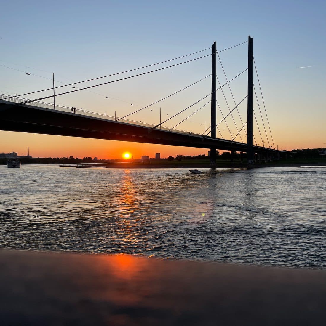 Eine Schrägseilbrücke als dunkle Silhouette über einem breiten Fluss, unter ihr geht die Sonne am Horizont unter und spiegelt sich orange im Wasser