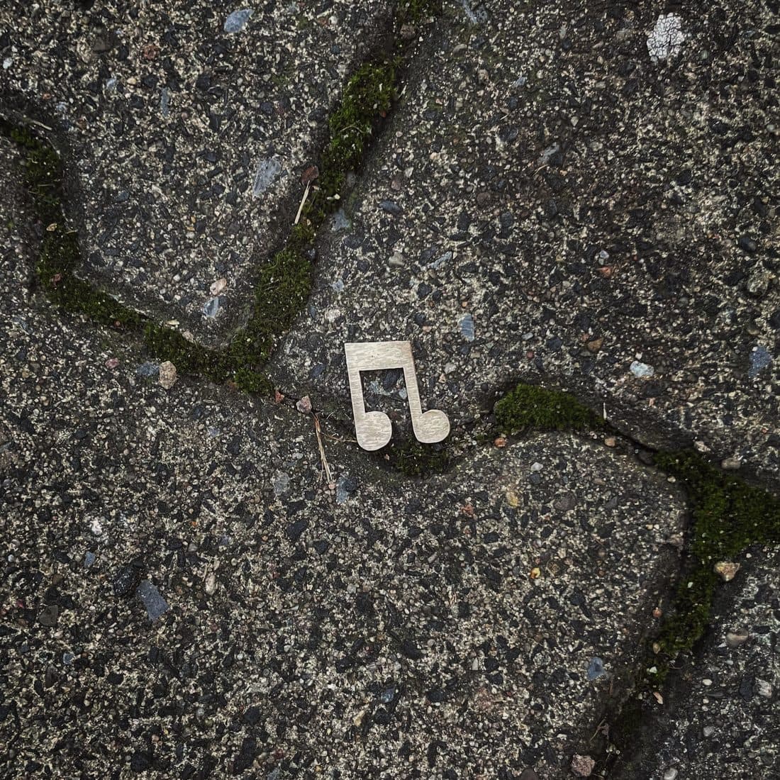 Ein flaches, hellbeiges Notensymbol aus zwei verbundenen Achtelnoten liegt auf grauem Verbundpflaster, in dessen geschwungenen Fugen Moos wächst