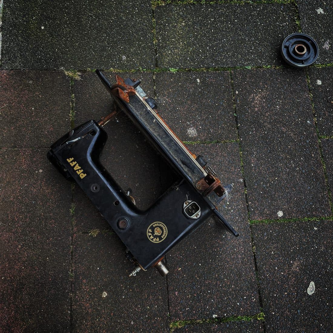 Das schwarze Gehäuse einer alten Pfaff-Nähmaschine mit goldenem Schriftzug und rostigen Teilen liegt auf dunklen Gehwegplatten, daneben ein loses rundes Teil