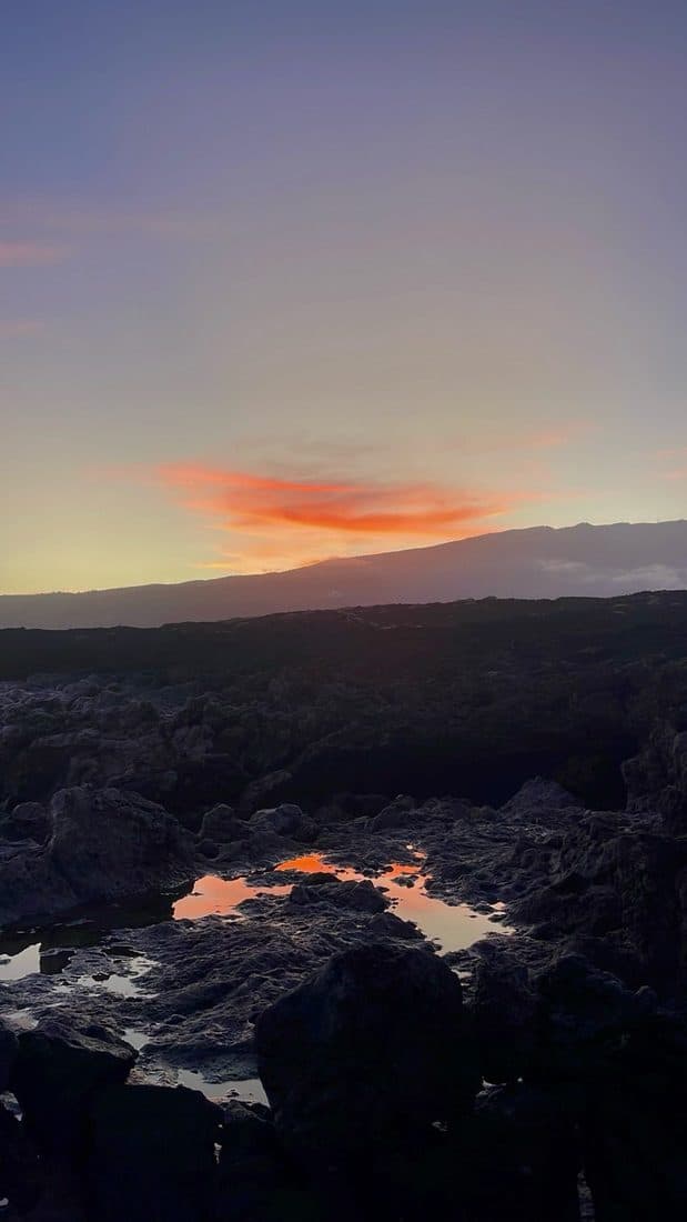 Über einem dunklen Lavafeld liegt ein orange leuchtender Wolkenstreifen am violetten Himmel; Wasserpfützen zwischen den Felsen spiegeln das Licht, hinten ein Bergmassiv