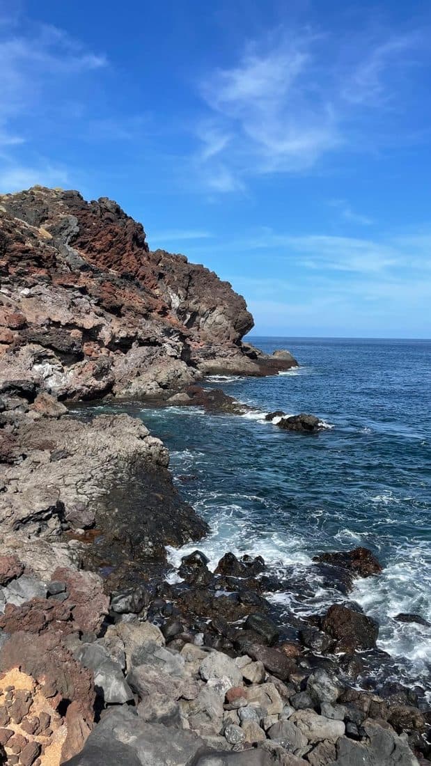 Eine Küste aus rotbraunem und grauem Lavagestein, davor tiefblaues Meer mit weißer Brandung, oben ein klarer Himmel