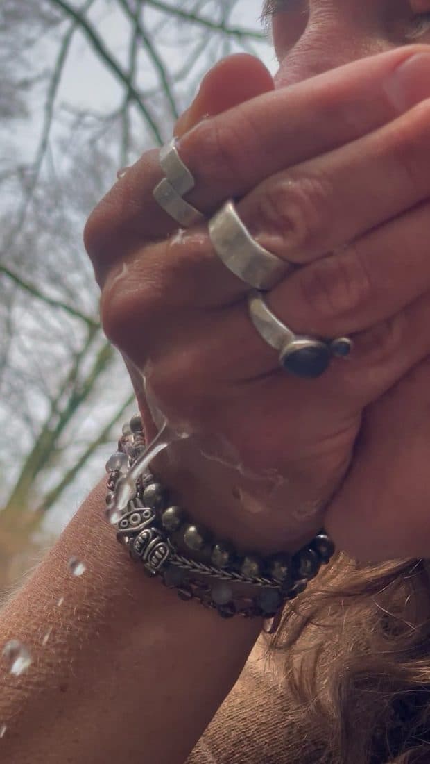 Nahaufnahme einer Hand mit vier breiten Silberringen und mehreren Perlenarmbändern, über die Wasser läuft, im Hintergrund kahle Zweige