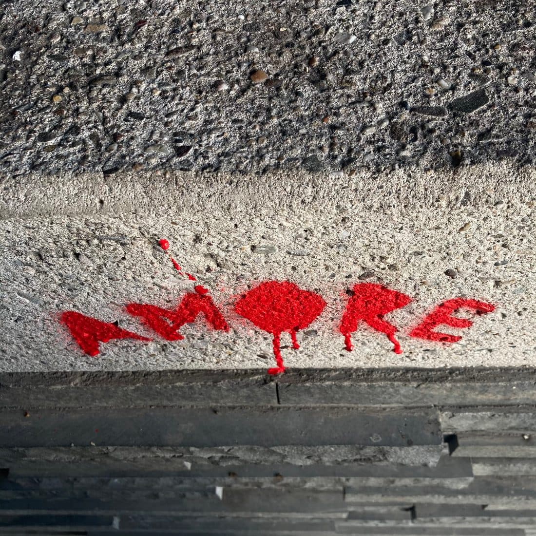 Das Wort AMORE in roter Sprühfarbe auf einer rauen Betonkante, das O als ausgefüllter Fleck mit herablaufenden Tropfen, darunter dunkle, geschichtete Steinplatten