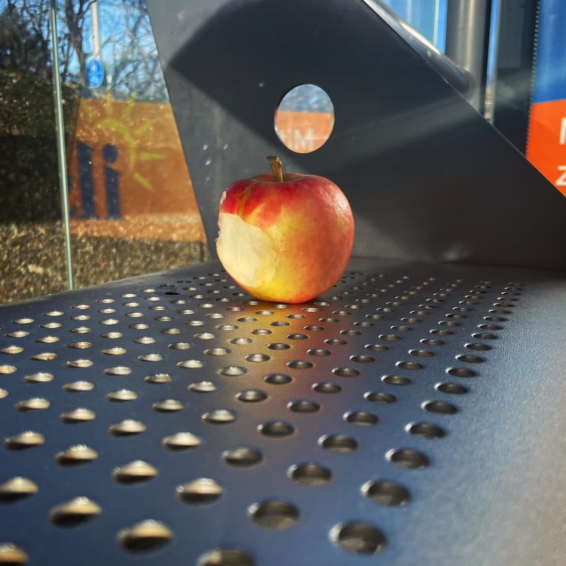 Ein angebissener rot-gelber Apfel steht in der Sonne auf einer gelochten, dunkelgrauen Metallbank, deren Lochränder hell aufblitzen, dahinter eine Glasscheibe