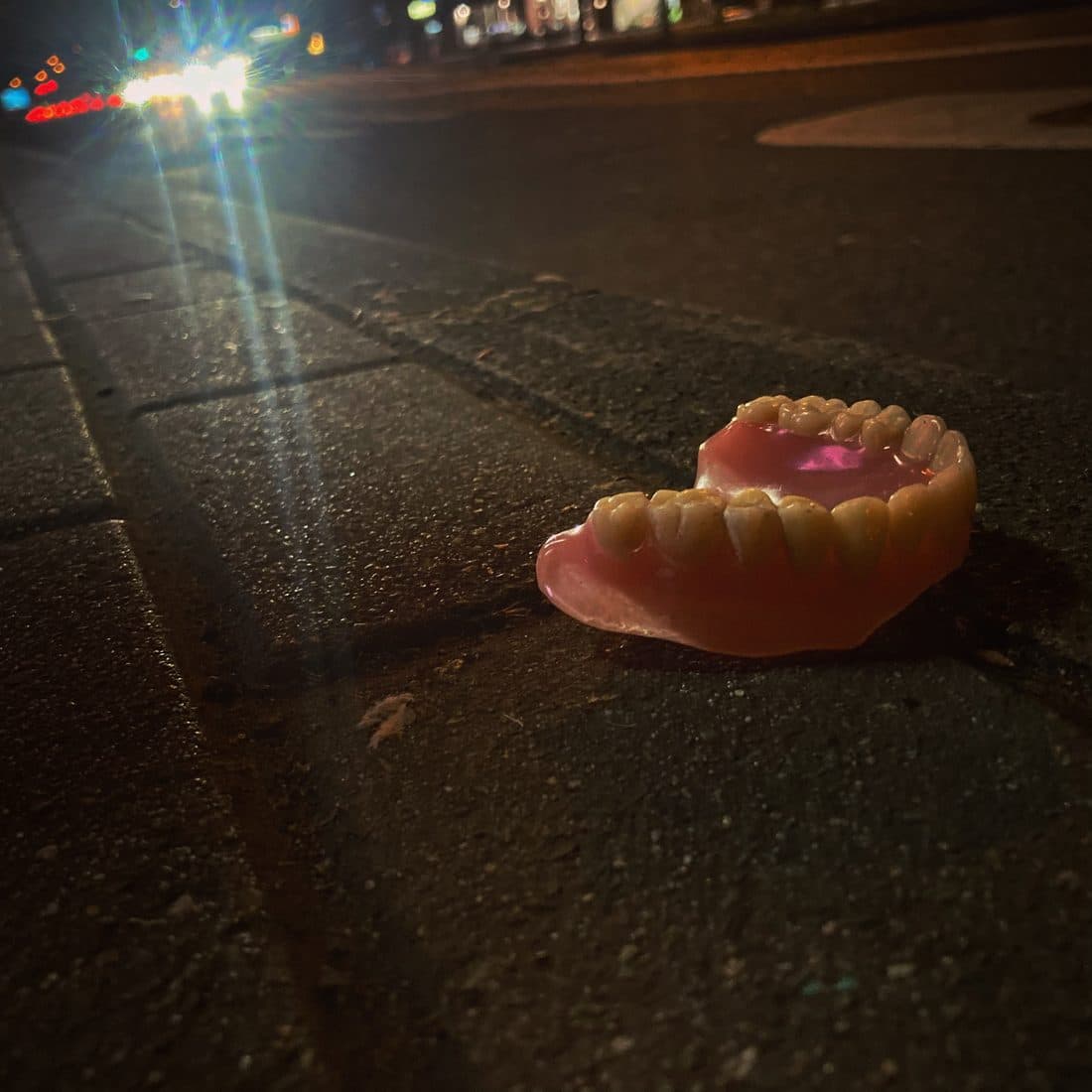 Eine Zahnprothese liegt nachts auf dem Pflaster am Straßenrand, dahinter das grelle Licht von Autoscheinwerfern und rote Rücklichter auf der dunklen Straße
