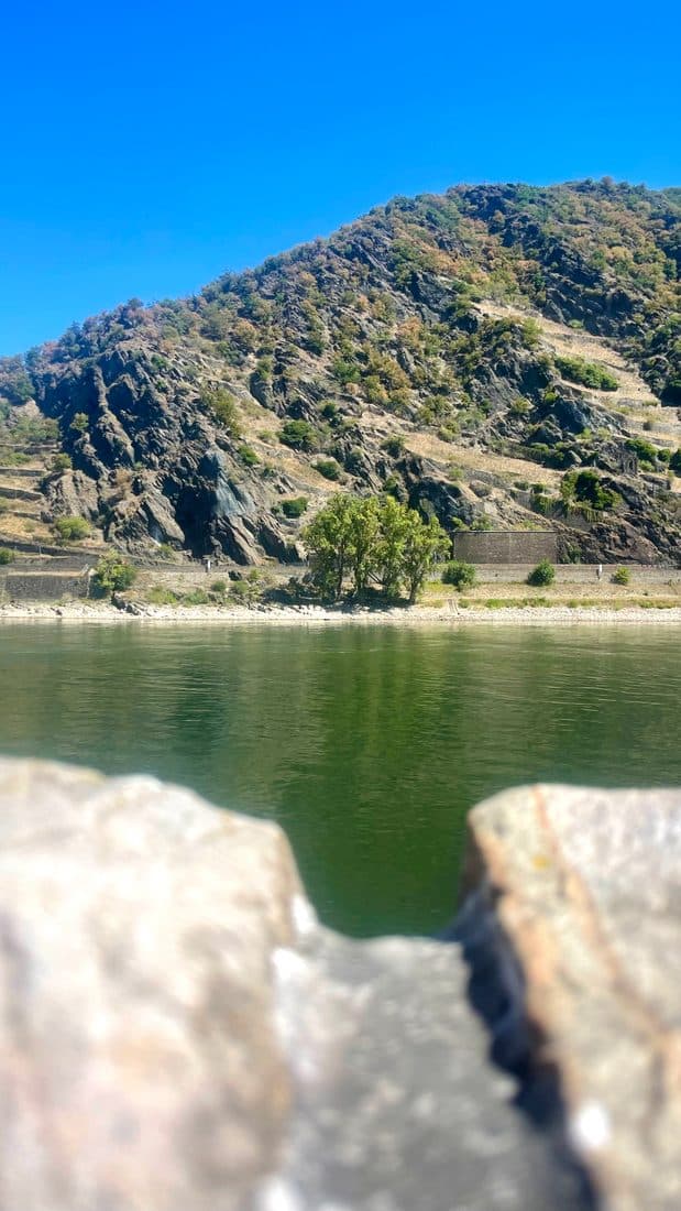 Der grüne Rhein und ein steiler Hang mit dunklem Fels und Büschen unter tiefblauem Himmel, gesehen durch eine Lücke zwischen zwei unscharfen Felsbrocken