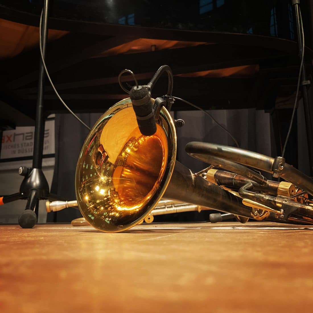 Ein Blechblasinstrument liegt auf einem warm beleuchteten Bühnenboden aus Holz, im glänzenden Schalltrichter spiegeln sich Lichter, am Rand ist ein Mikrofon befestigt
