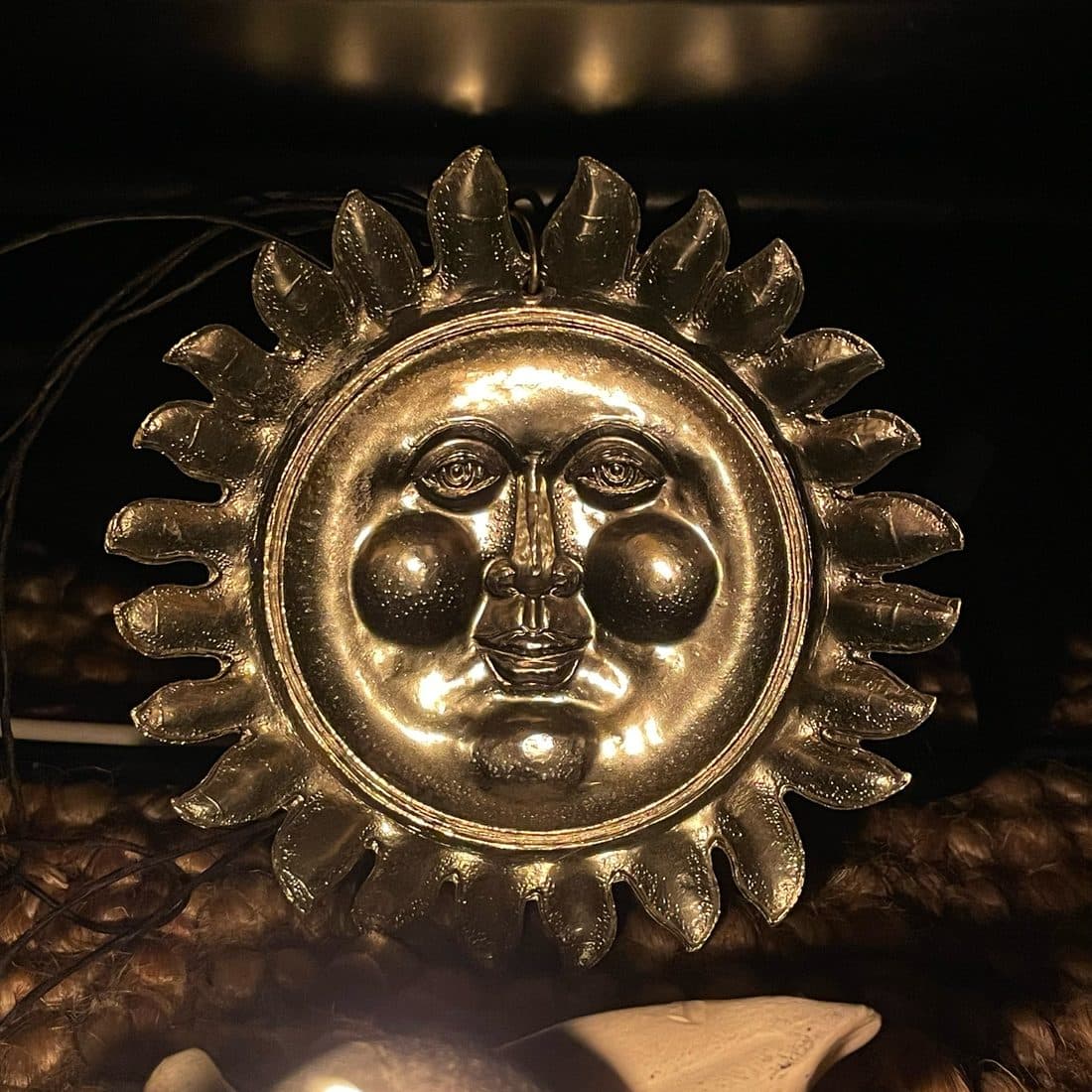 Eine metallene Sonne mit Gesicht und geschwungenen Strahlen, warm angestrahlt vor dunklem Hintergrund, darunter ein geflochtener Untergrund