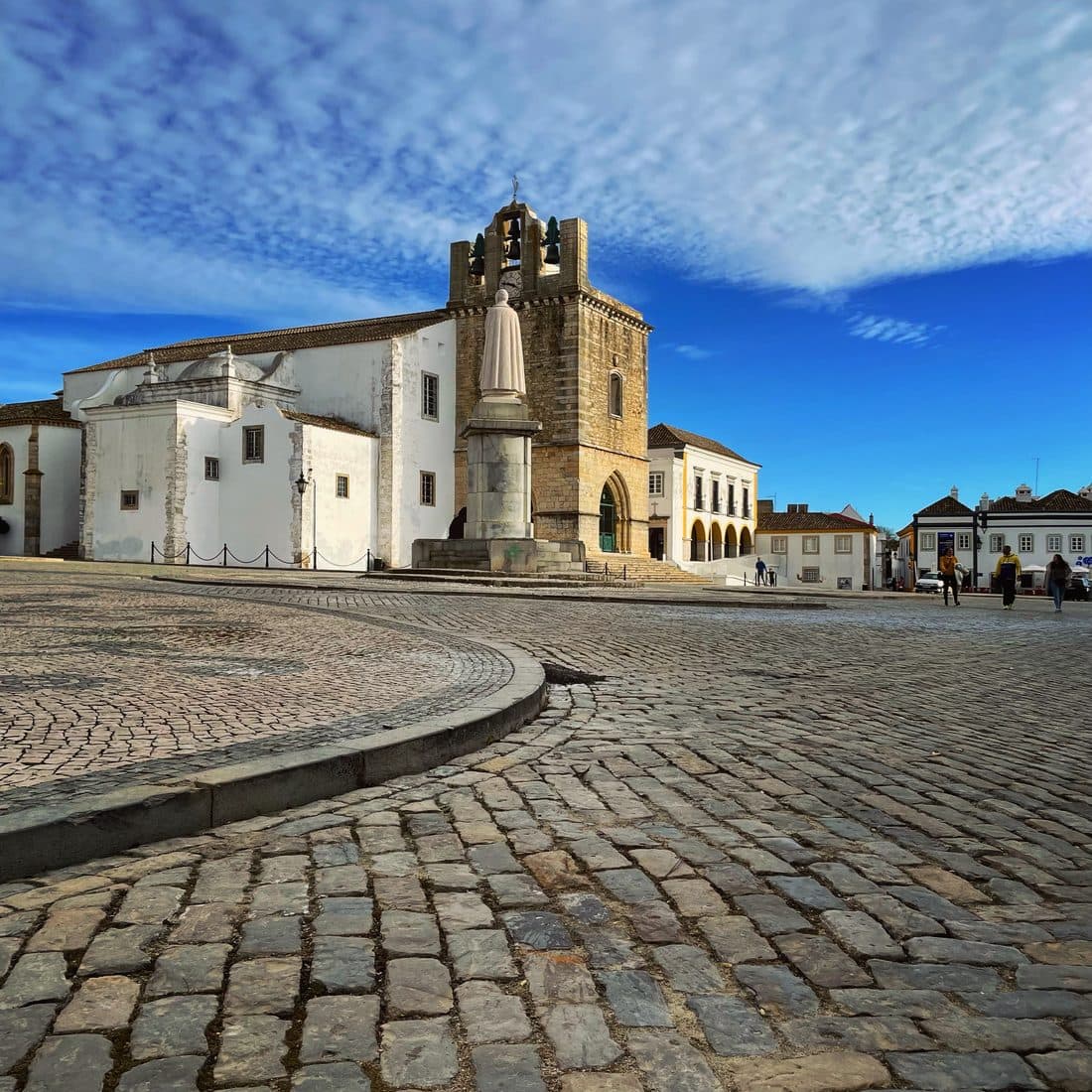 Die Kathedrale von Faro mit weißem Kirchenbau und steinernem Glockenturm, davor eine helle Statue auf hohem Sockel und ein weiter Platz aus Kopfsteinpflaster unter feinen Schäfchenwolken
