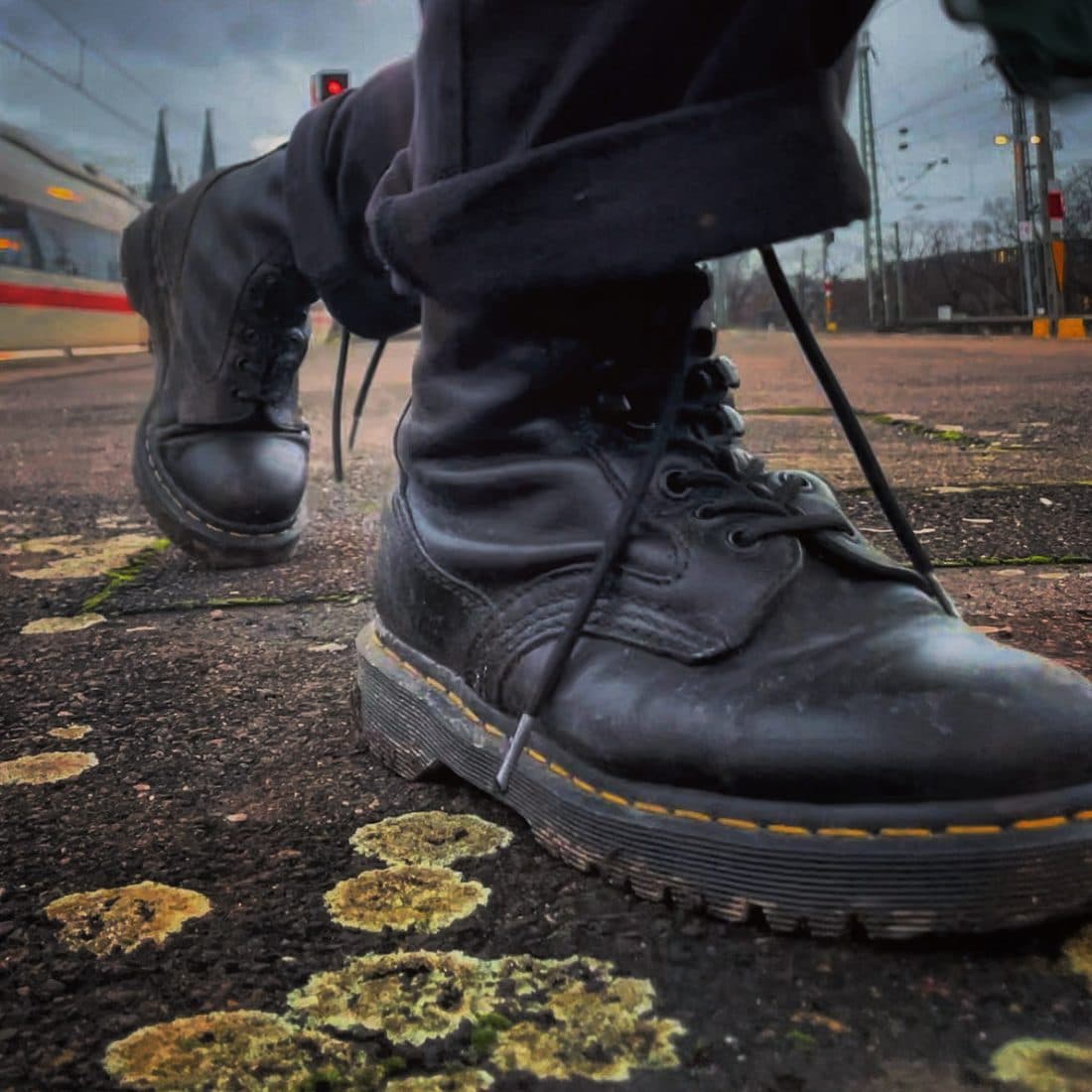 Die Füße einer Person in schwarzen Schnürstiefeln mit gelber Naht, mitten im Schritt aus Bodenhöhe gesehen, auf Asphalt mit gelben Flechten, dahinter ein Zug und zwei Kirchturmspitzen