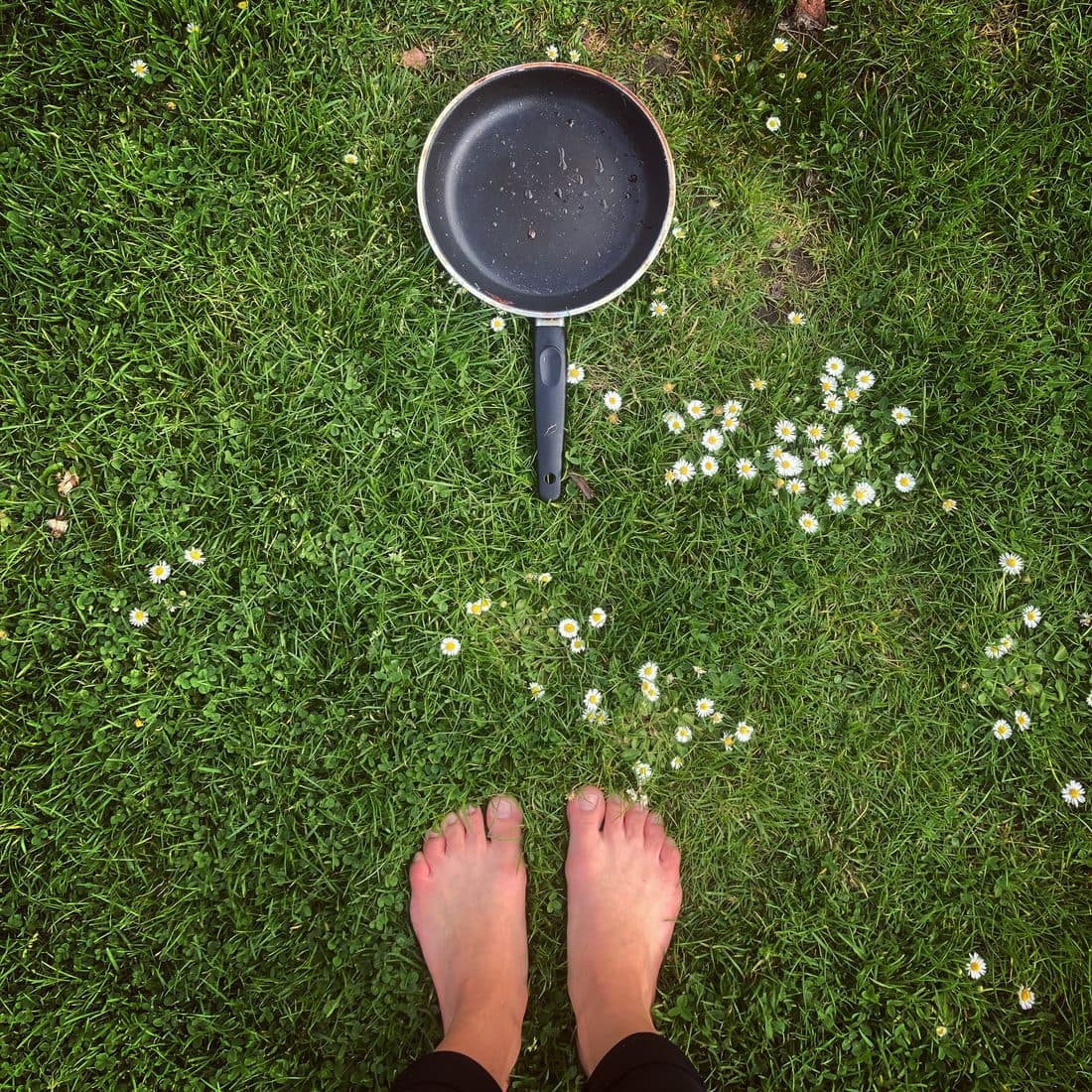 Von oben gesehen stehen zwei nackte Füße auf einem Rasen mit Gänseblümchen, vor ihnen liegt eine schwarze Bratpfanne mit Wassertropfen im Gras