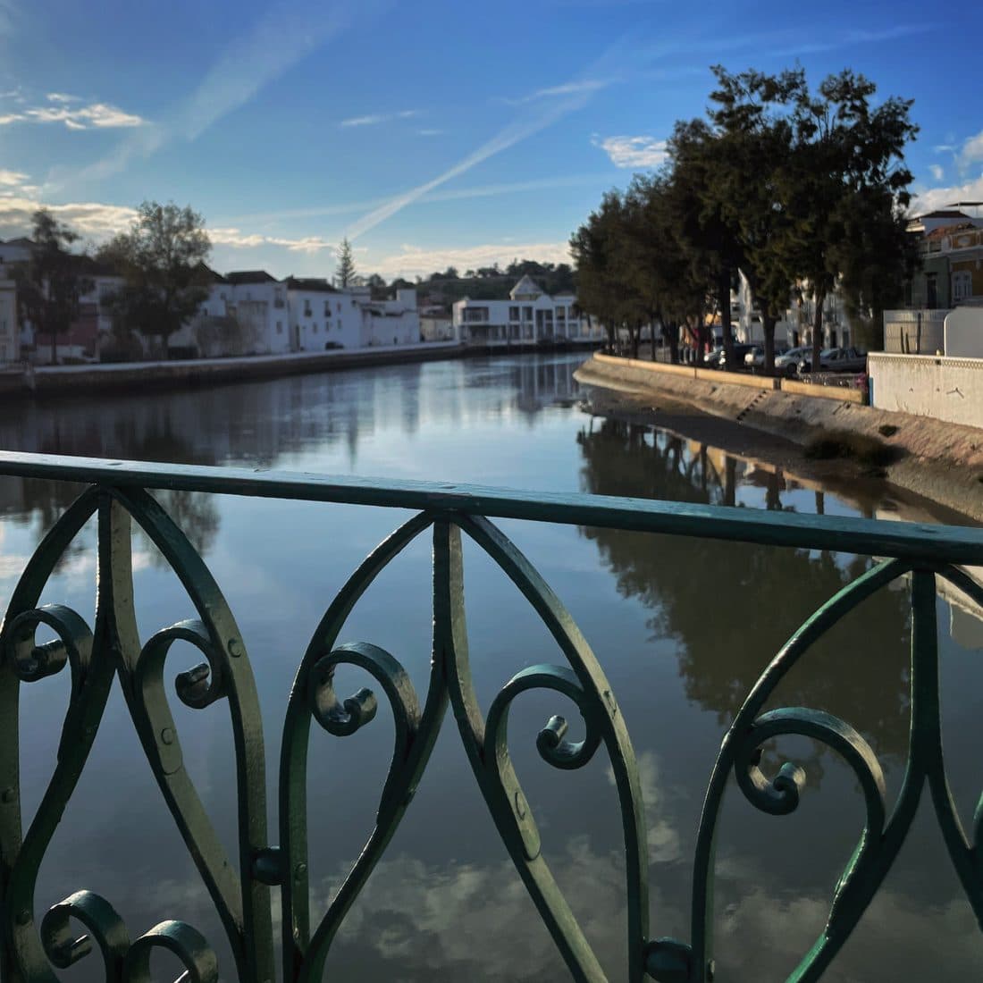 Ein grünes, verschnörkeltes Eisengeländer vor einem ruhigen Fluss, in dem sich der blaue Himmel, Bäume und weiße Häuser am Ufer spiegeln
