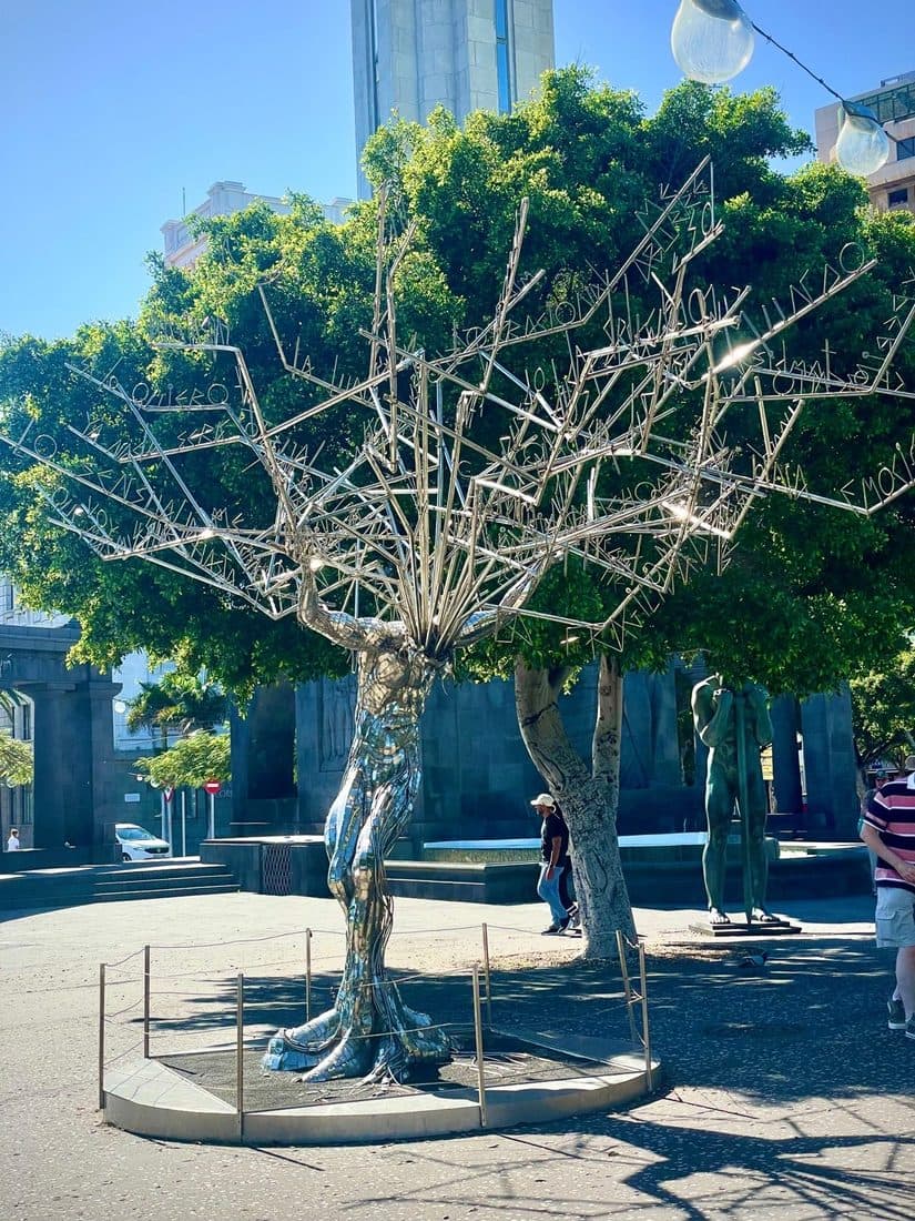 Eine Skulptur aus glänzenden Metallstäben in Form eines Baumes, dessen Äste aus Buchstaben bestehen, auf einem sonnigen Platz vor Bäumen