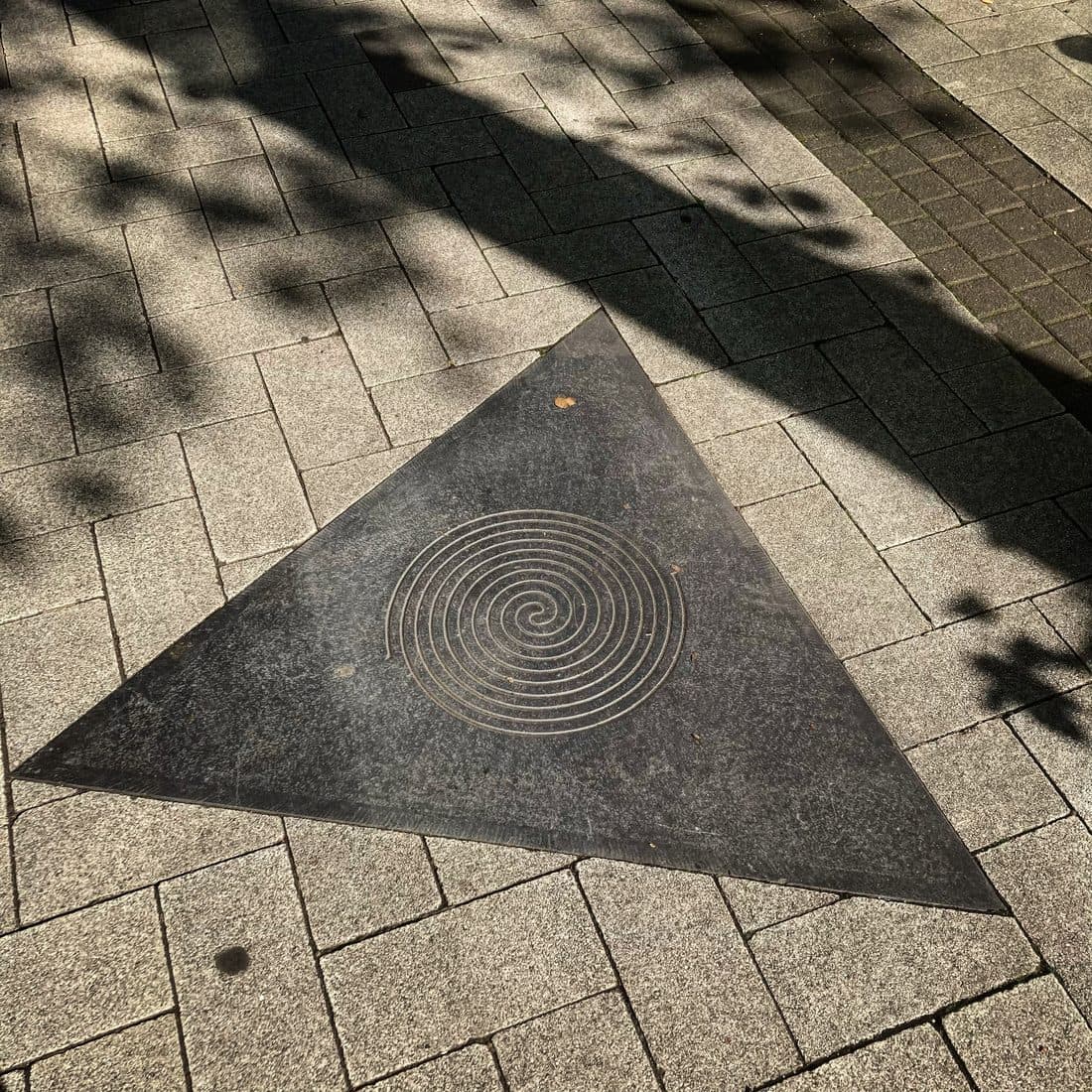 Eine dreieckige, dunkle Steinplatte mit eingelassener heller Spirale im grauen Pflaster, darüber die Schatten von Zweigen und Blättern im Sonnenlicht
