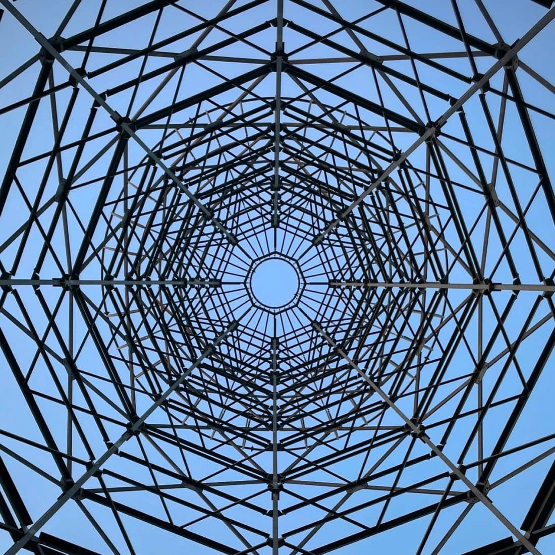 Blick von unten in einen Stahlgittermast: verstrebte Achtecke staffeln sich nach oben, in der Mitte ein helles Loch, dahinter blauer Himmel