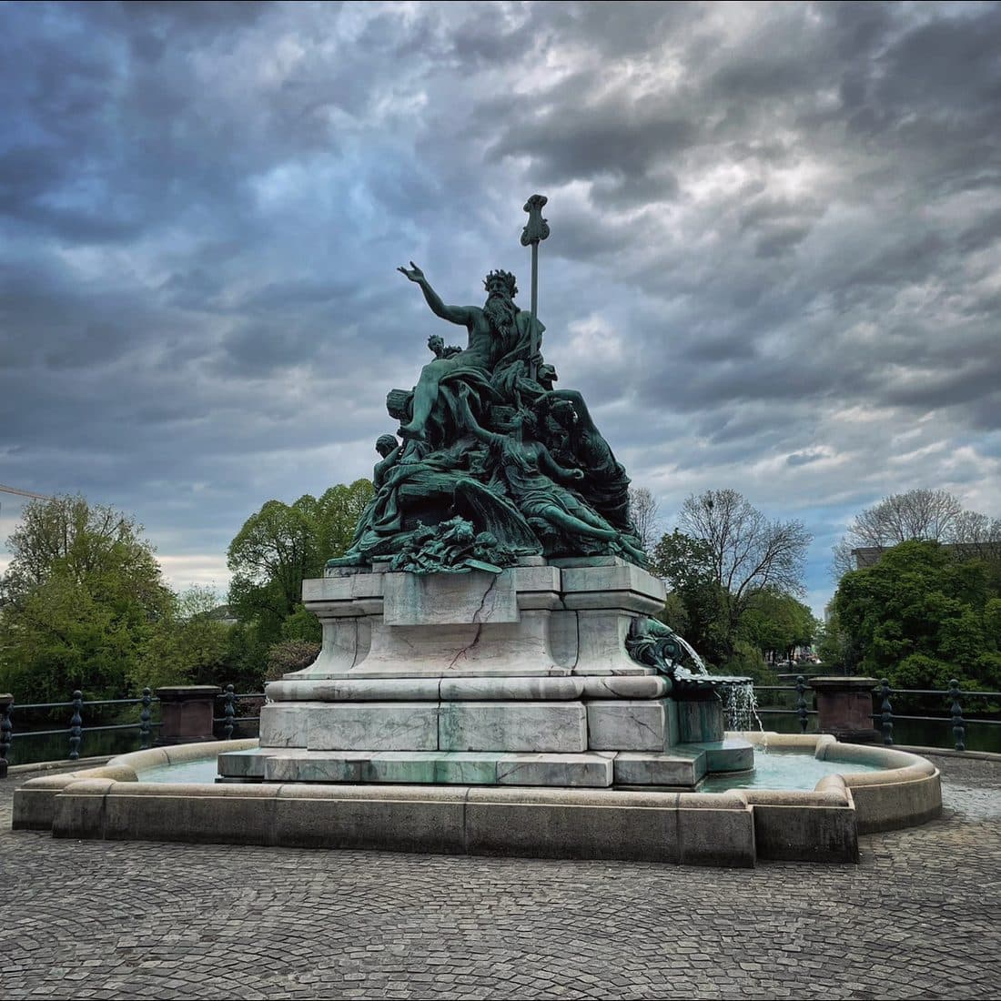 Eine grün patinierte Bronzegruppe mit einer bärtigen, gekrönten Figur, die einen Arm hebt, auf dem hellen Steinsockel eines runden Brunnens unter dicht bewölktem Himmel
