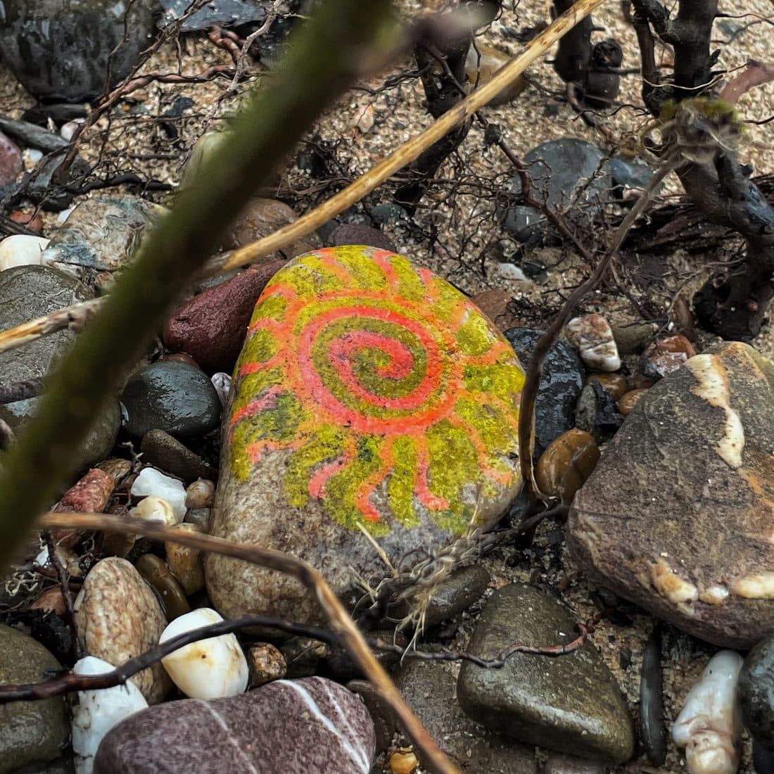 Ein ovaler Stein, auf den eine Sonne mit Spirale in Orange und Gelbgrün gemalt ist, liegt zwischen glatten Kieseln, Sand und trockenen Wurzeln