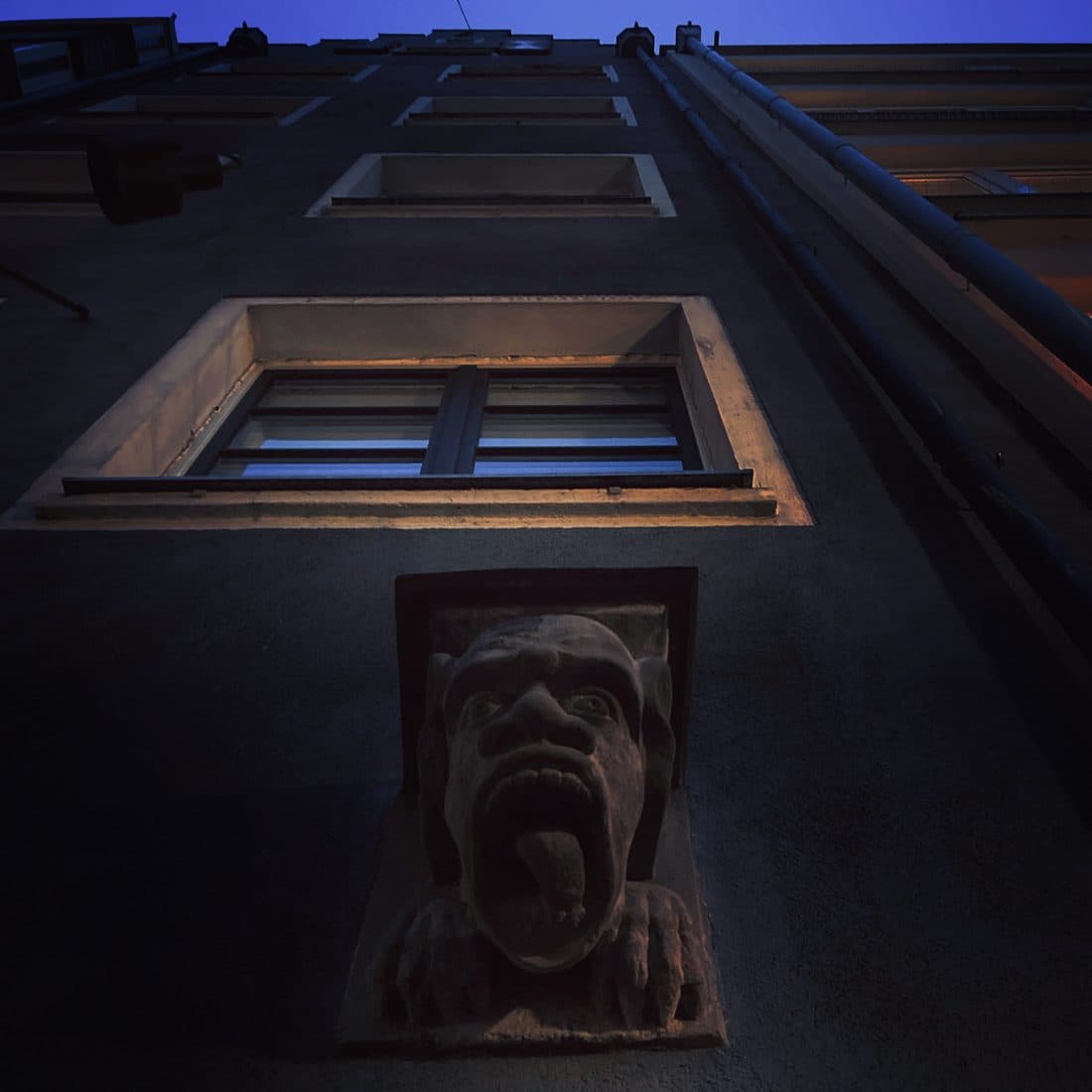 Eine steinerne Fratze mit herausgestreckter Zunge an einer dunklen Hausfassade, von unten gesehen, darüber ein warm angestrahltes Fenster und der tiefblaue Abendhimmel