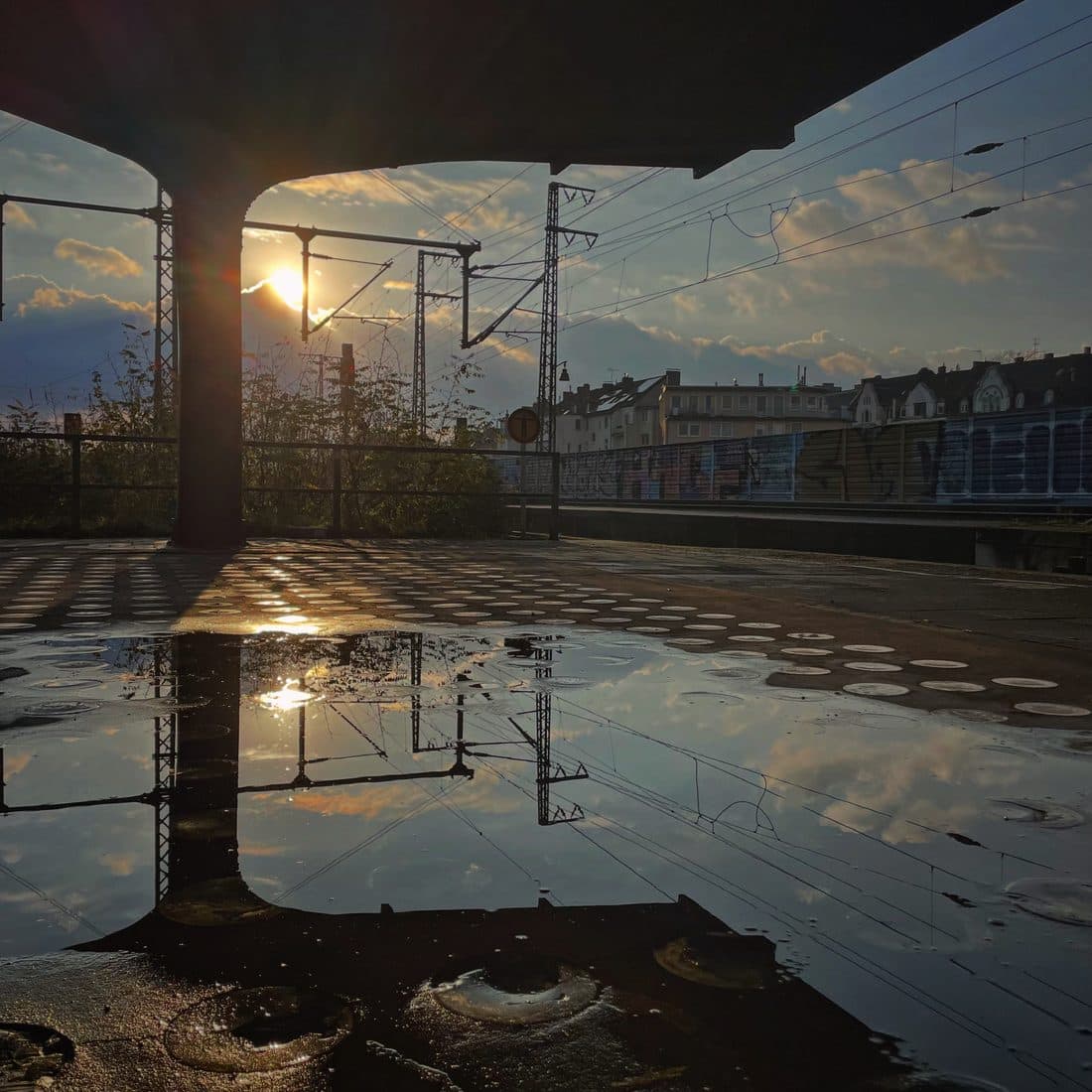 Eine große Pfütze unter einem Bahnsteigdach spiegelt Sonne, Wolken und Oberleitungsmasten, dahinter Häuser und eine mit Graffiti besprühte Lärmschutzwand im Gegenlicht