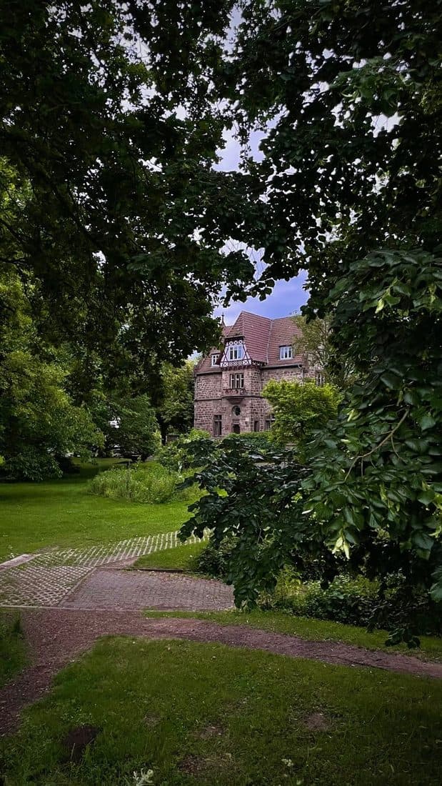Durch dichtes Laub hindurch ein Haus aus rotem Sandstein mit Fachwerkgiebel und Ziegeldach, davor eine Wiese mit Weg